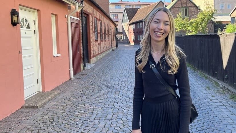 Hanna Bergenholtz står på en kullerstensgata omgiven av färgglada hus och staket i en solig stadsmiljö. Hon bär en mörk outfit och har långt blont hår.