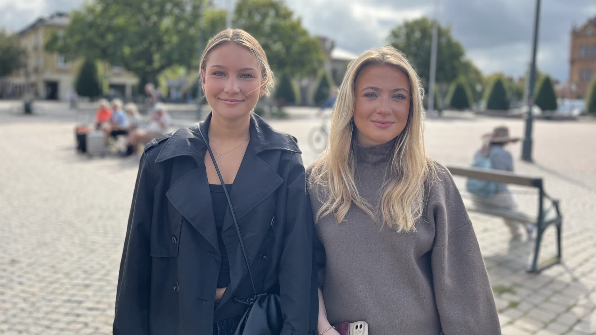 Två kvinnor, Agnes Aarnes och Agnes Åkesson, står utomhus med ett torg i bakgrunden. Träd och en byggnad syns i fjärran under en molnig himmel.