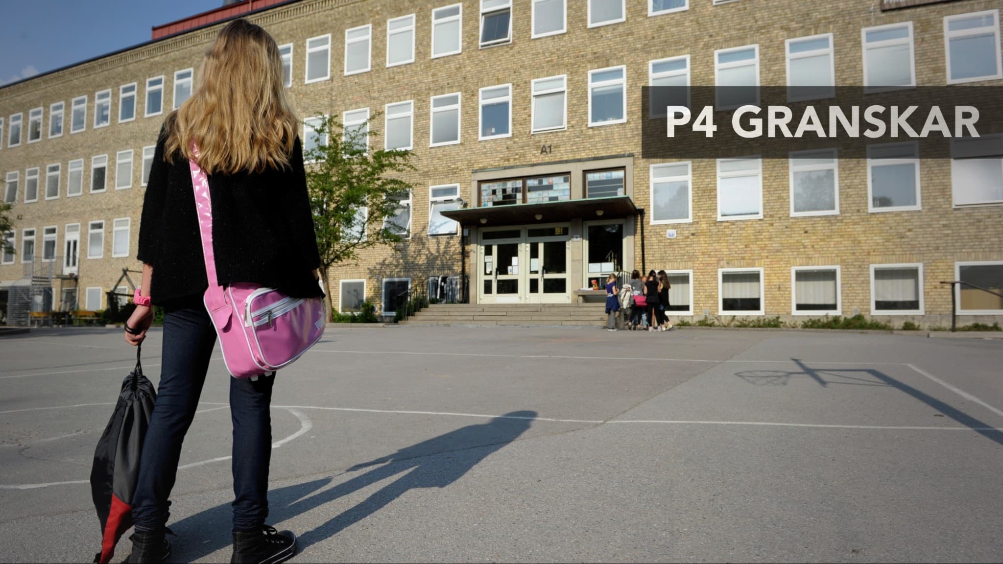 En flicka med ryggsäck och rosa väska står på en skolgård framför en skolbyggnad med gula tegelväggar. Skylten 'P4 Granskar' syns i högra hörnet av bilden.