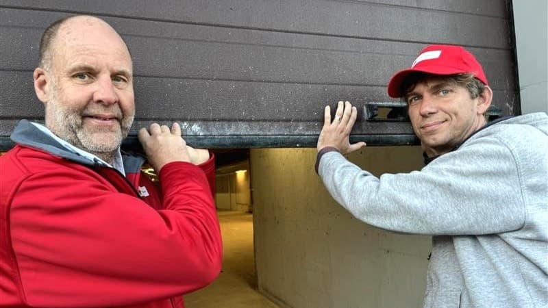 Två personer öppnar en garageport i en industriell miljö. Den ena bär en röd jacka och den andra en grå hoodie med keps.