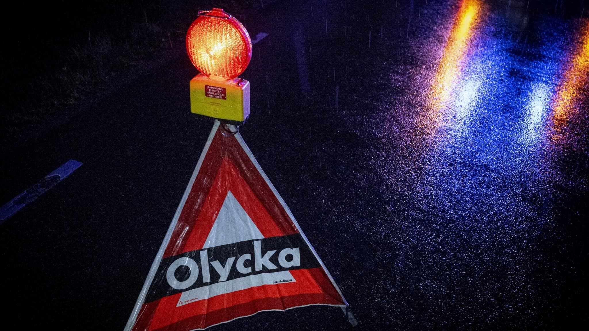 En olyckspyramid med texten 'Olycka' står på en avstängd väg i mörker, upplyst av blåljus och reflektioner på den våta asfalten. 