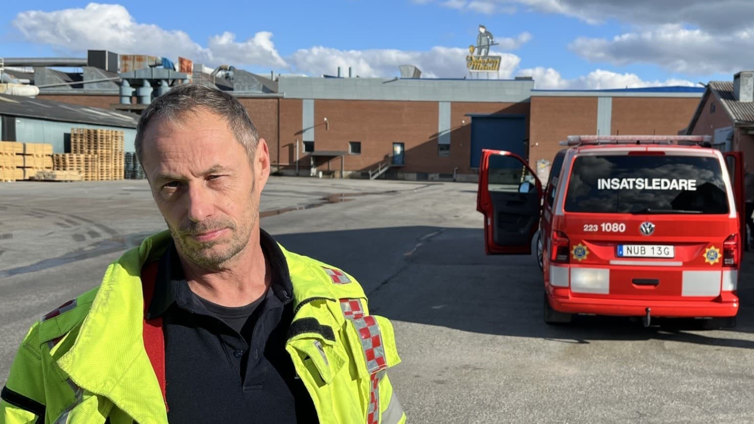 En brandman i gul skyddsutrustning står framför en röd räddningstjänstbil på en industriell gårdsplan med lagerbyggnader i bakgrunden. Solen skiner och himlen är blå med några moln.
