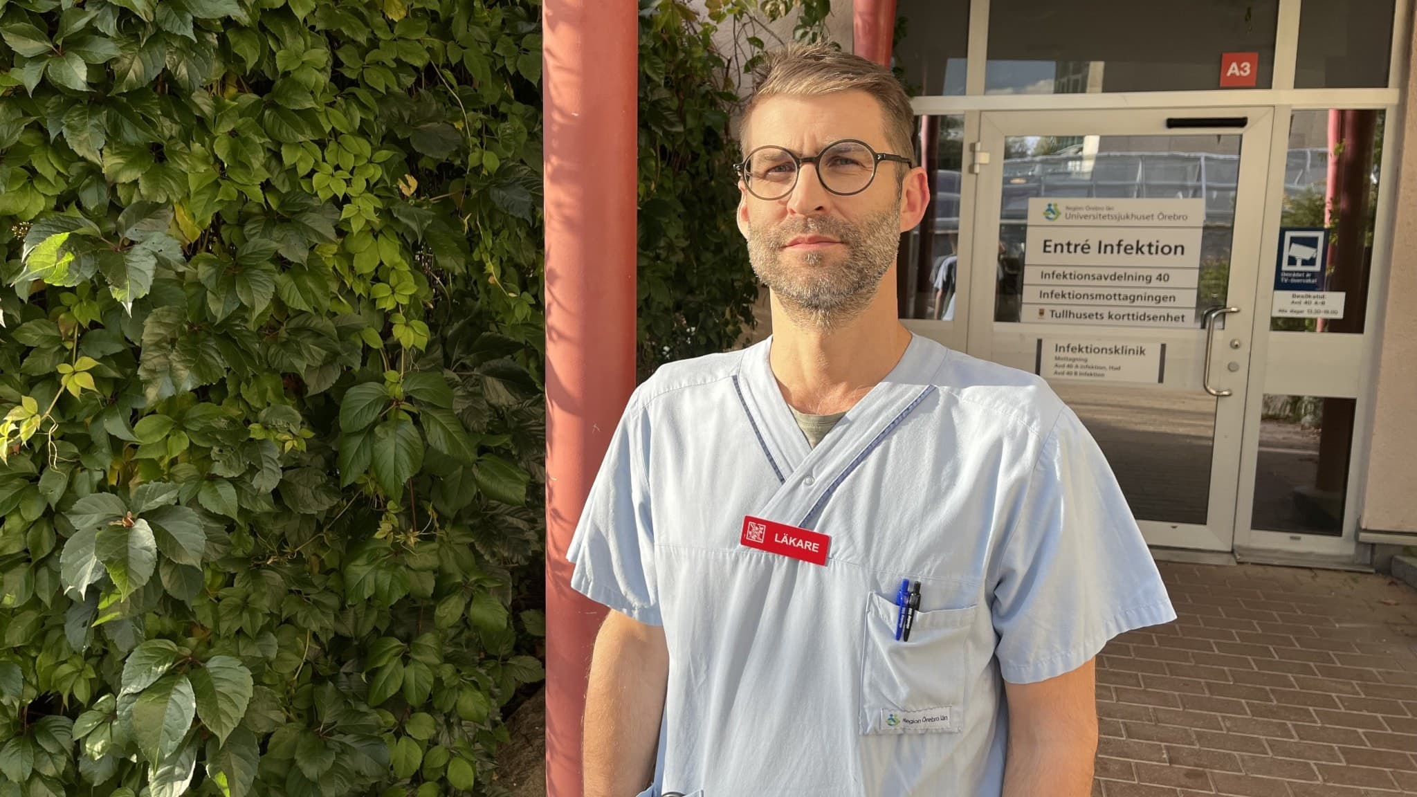 Martin Widlund, läkare klädd i ljusblå arbetskläder, står utomhus framför en grön växtvägg och en glasentré med skyltar. Han har en namnbricka och pennor i bröstfickan.