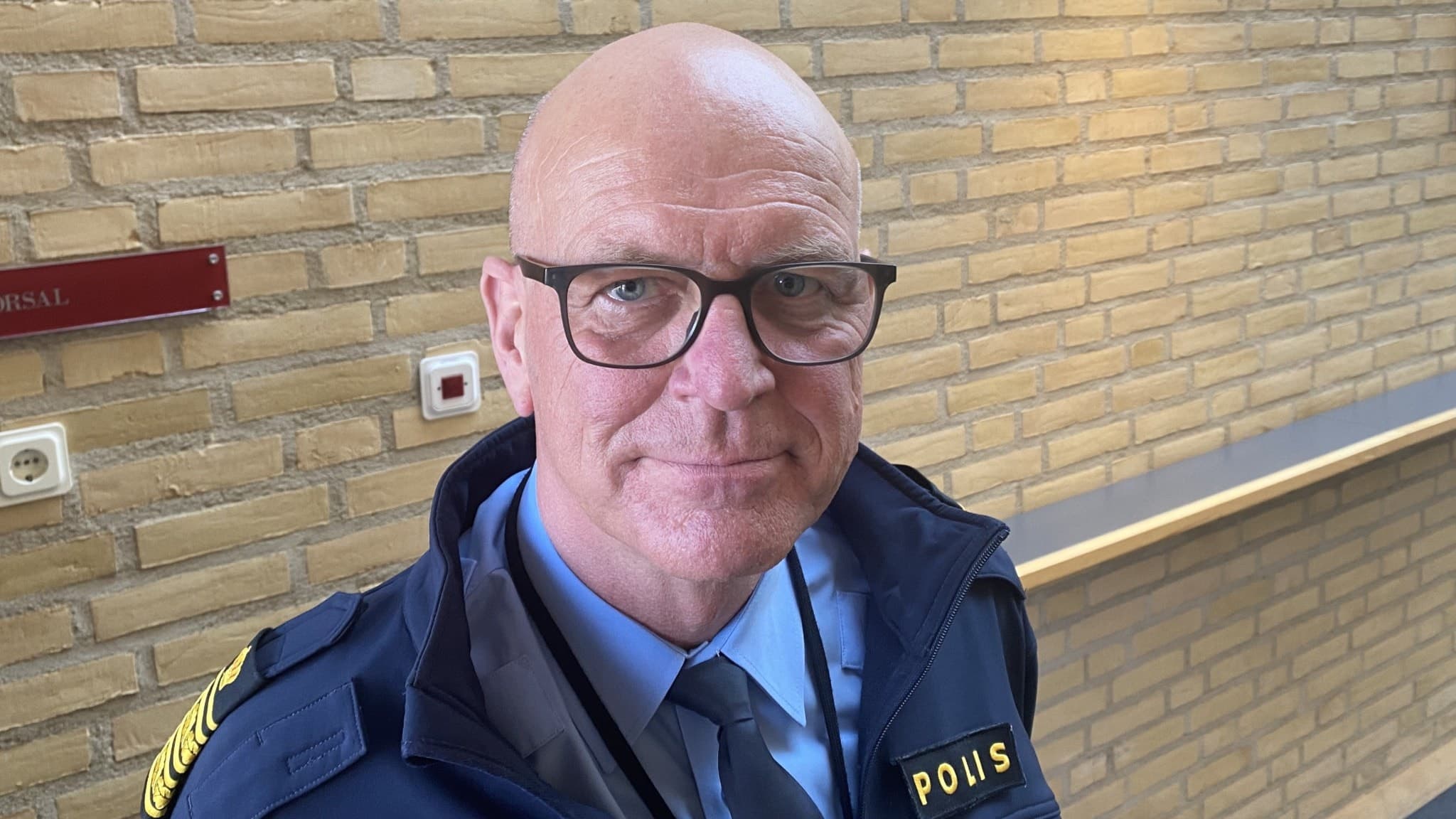 Erik Nord, polis, i uniform framför en gul tegelvägg med en röd namnskylt i bakgrunden. Han syns stående inomhus, klädd i blå skjorta och mörkblå jacka med texten 'Polis'.