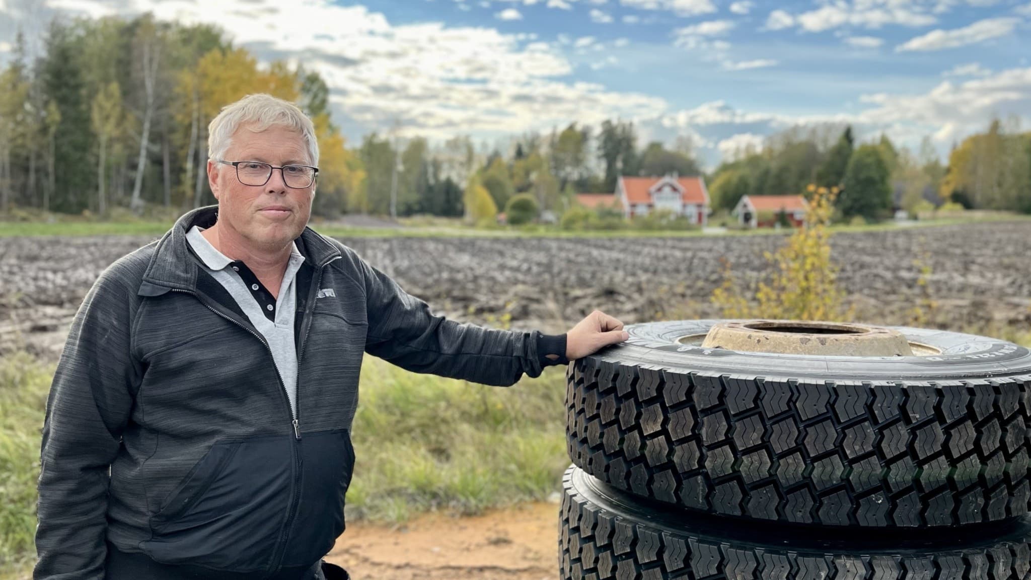 Morgan Jakobsson står vid två stora traktordäck på en landsbygd med ett plöjt fält i bakgrunden och ett hus omgiven av träd. Himlen är klar med några moln.