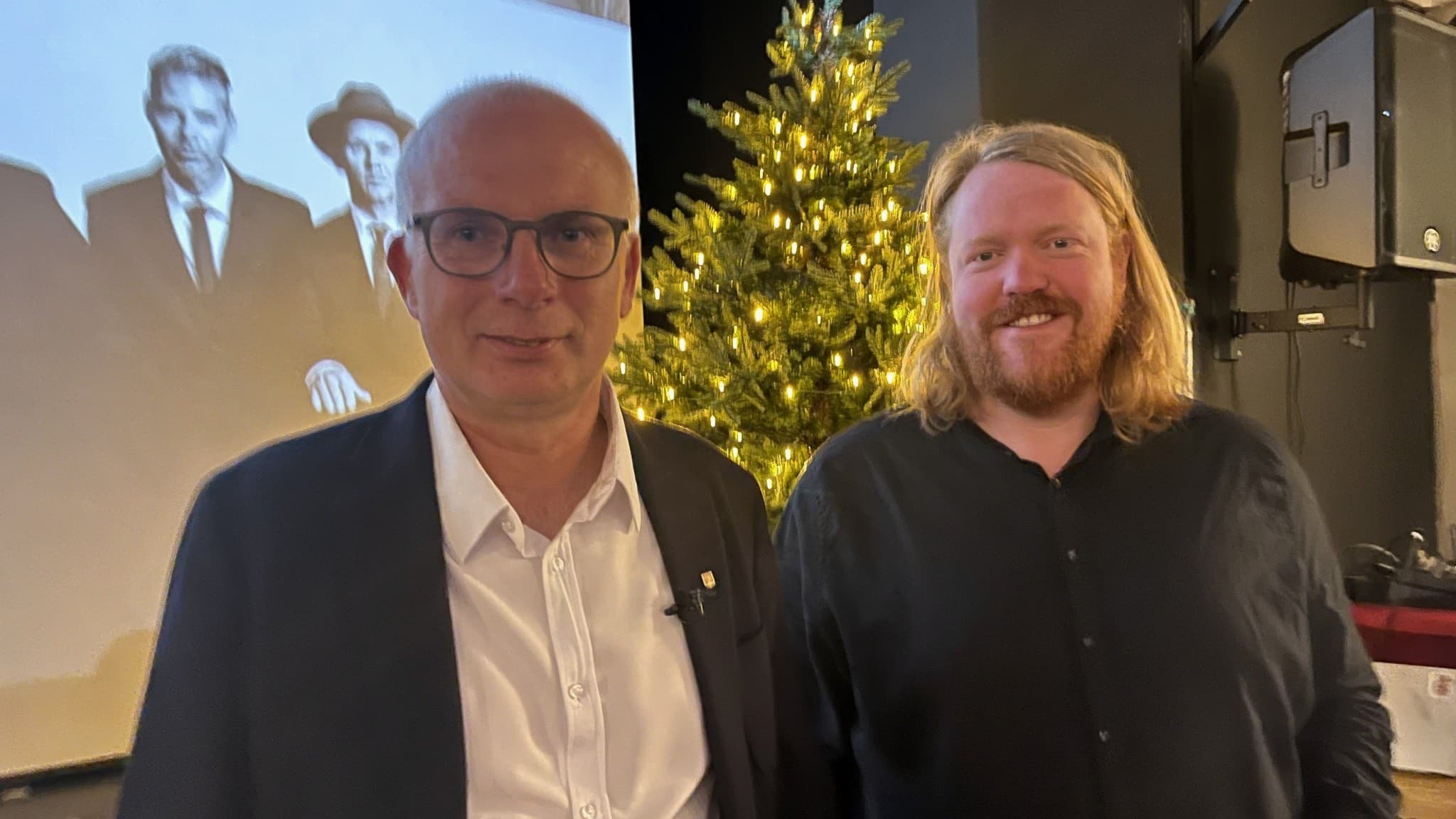 Henrik Arnevang och Martin Almgren står framför en julgran i en inomhusmiljö. I bakgrunden syns en projicerad bild med personer i kostymer.