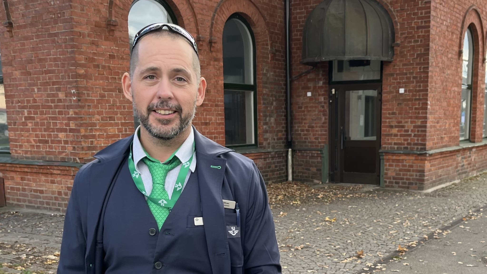 Mikael Freiholtz, lokförare, står utomhus framför en äldre tegelbyggnad med bågformade fönster och kullerstensmark. Han bär uniform med grön slips och har händerna i fickorna.