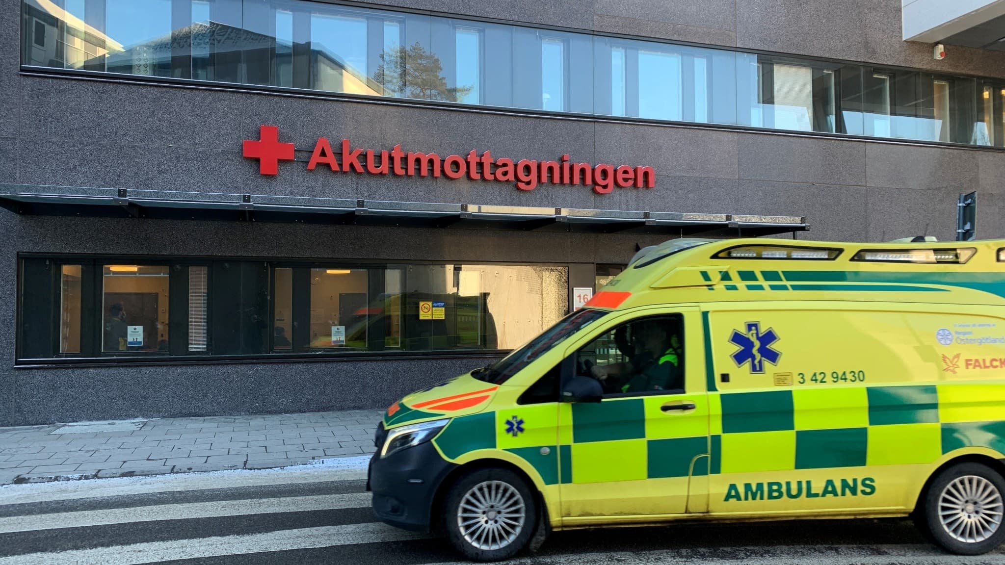 En gul ambulans står parkerad framför akutmottagningen vid Universitetssjukhuset i Linköping. Byggnaden har en grå fasad och röda bokstäver som markerar ingången.