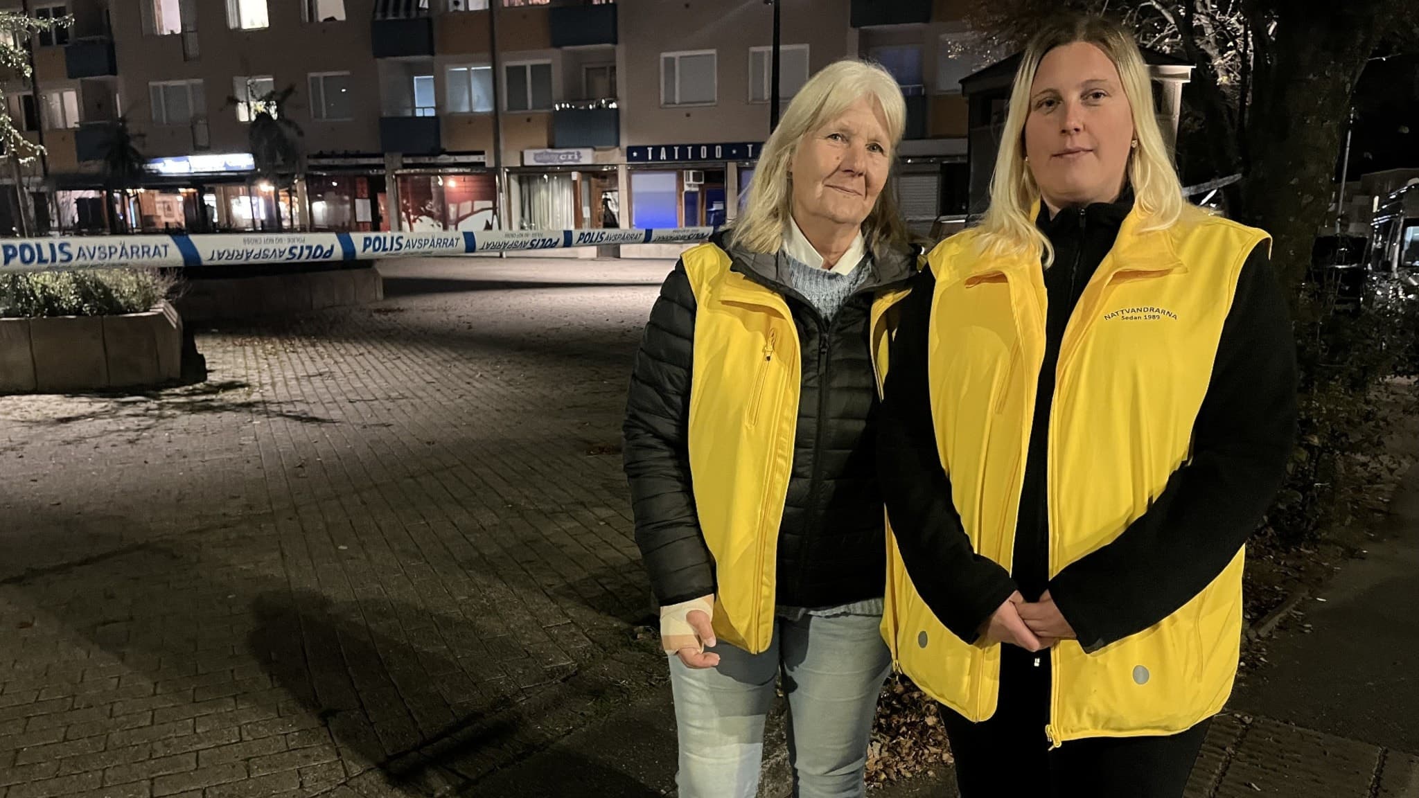Två personer i gula västar från nattvandrargruppen står vid en avspärrad plats utomhus, där polisens avspärrningsband syns i bakgrunden. Mörka byggnader och en torgmiljö omger området.