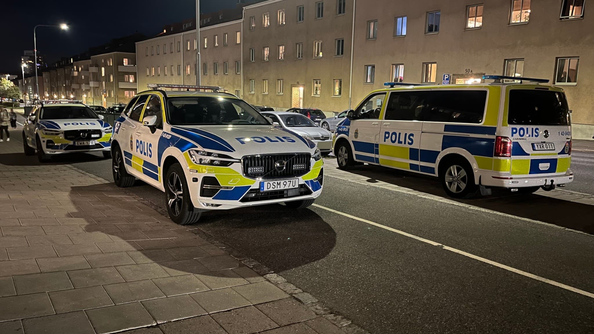 Flera polisfordon står parkerade längs en gata i ett bostadsområde under kvällstid. Polisens insats pågår, med fokus på trygghet och säkerhet efter en händelse i området.