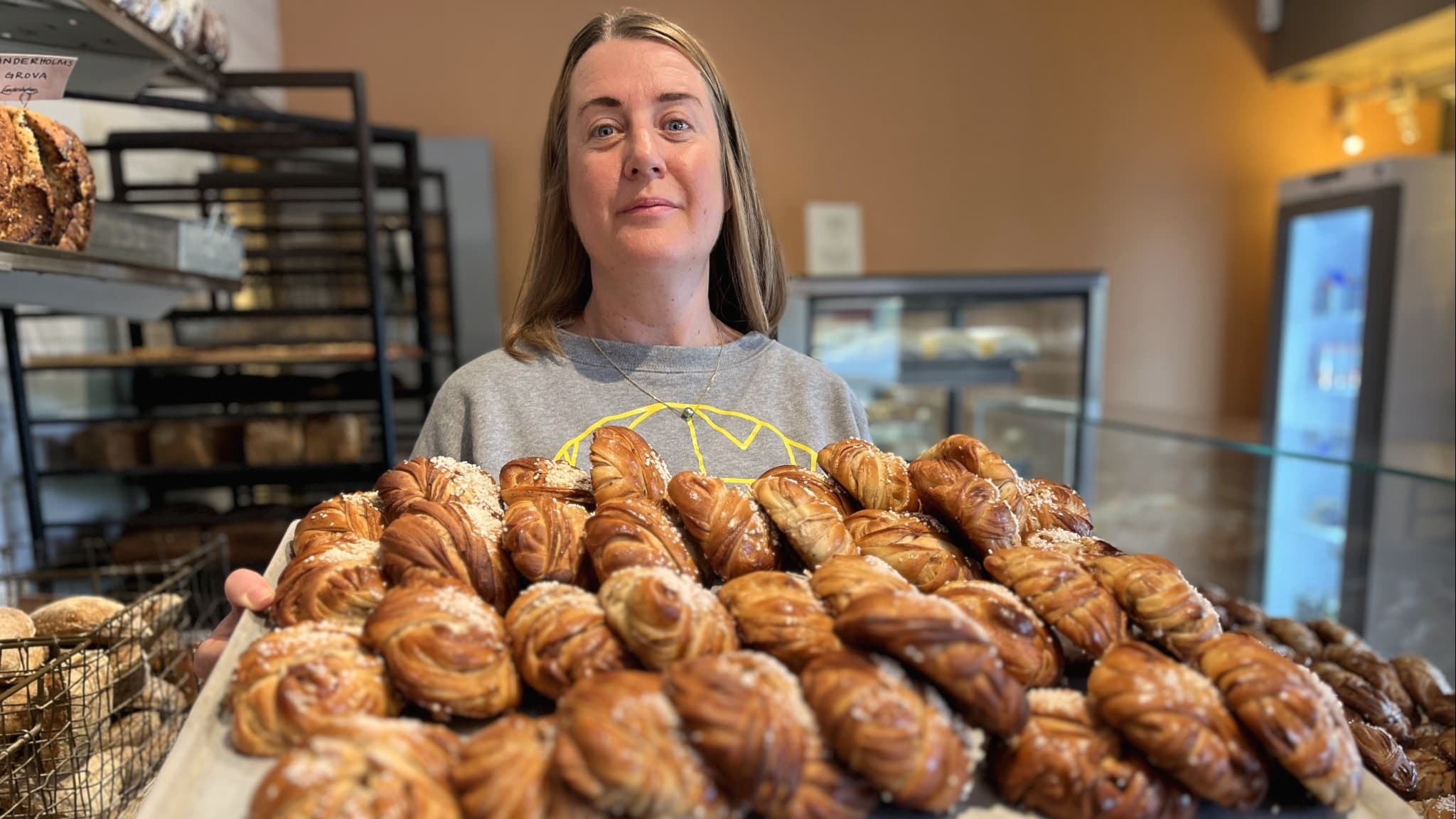 Sofia Landerholm, delägare i ett bageri i Norrköping, håller en bricka fylld med nygräddade kanelbullar. I bakgrunden syns hyllor med bröd och en kyl med bakverk.