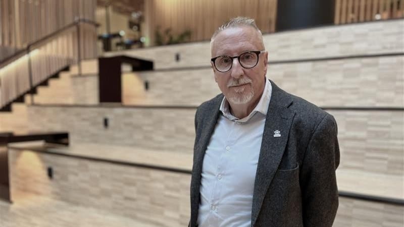 En man i grå kavaj och vit skjorta står i en modern byggnad med trappor och träpaneler, som kan vara en del av Högskolan. Miljön är ljus och inbjudande med en stilren design.