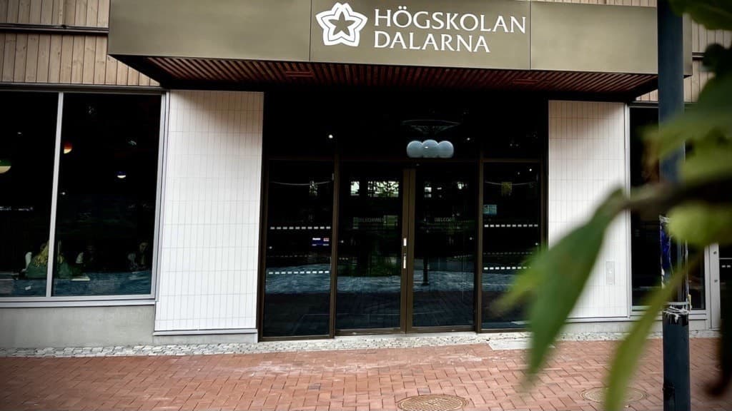 Entrén till Högskolan Dalarna med glasdörrar och en skylt med skolans namn och logotyp ovanför. En buske syns i förgrunden. Tegelmarken utanför skapar en välkomnande känsla.