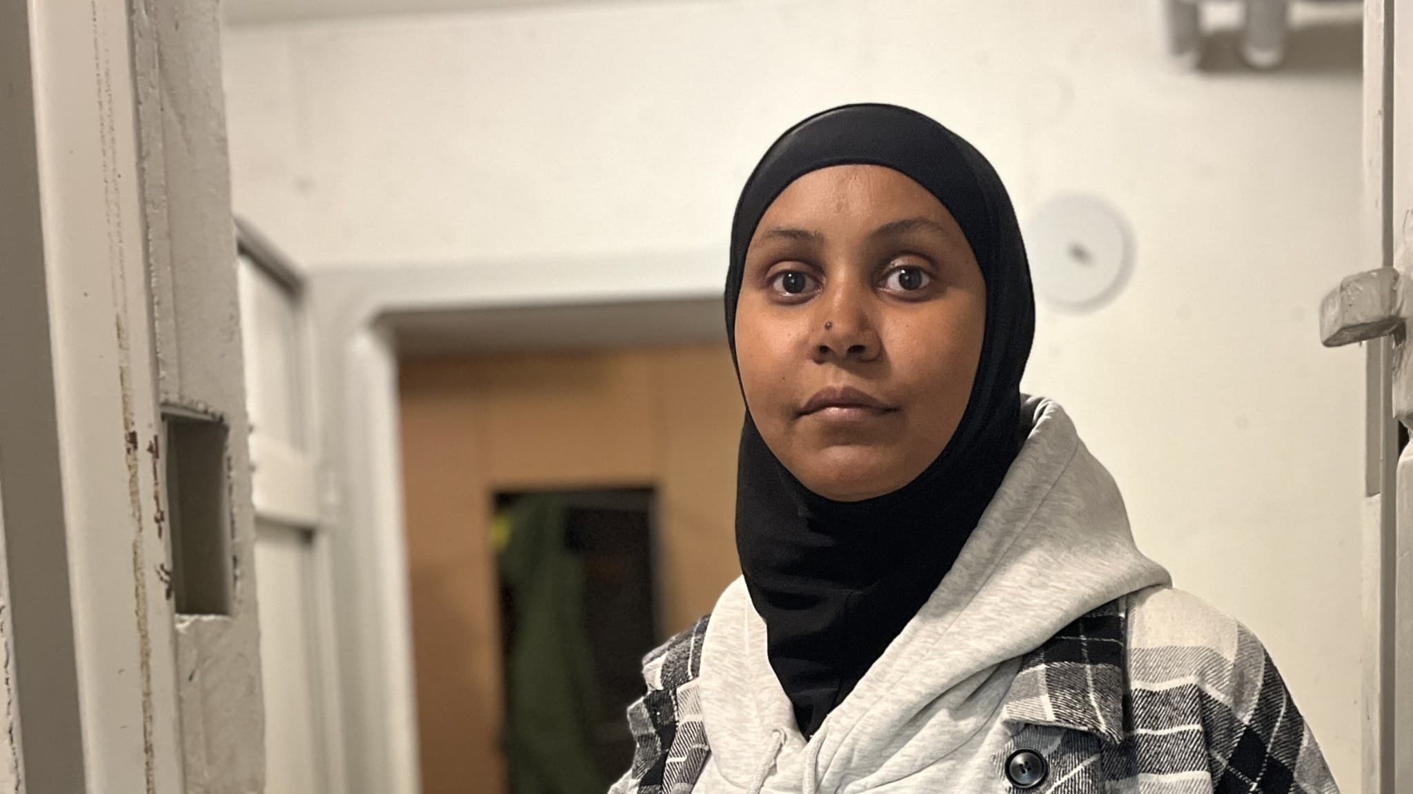 Sara Hussein står i ett skyddsrum som visas upp av Mora kommun. Hon bär en svart hijab, grå huvtröja och en rutig jacka. Miljön är enkel med vita väggar och en dörröppning i bakgrunden.