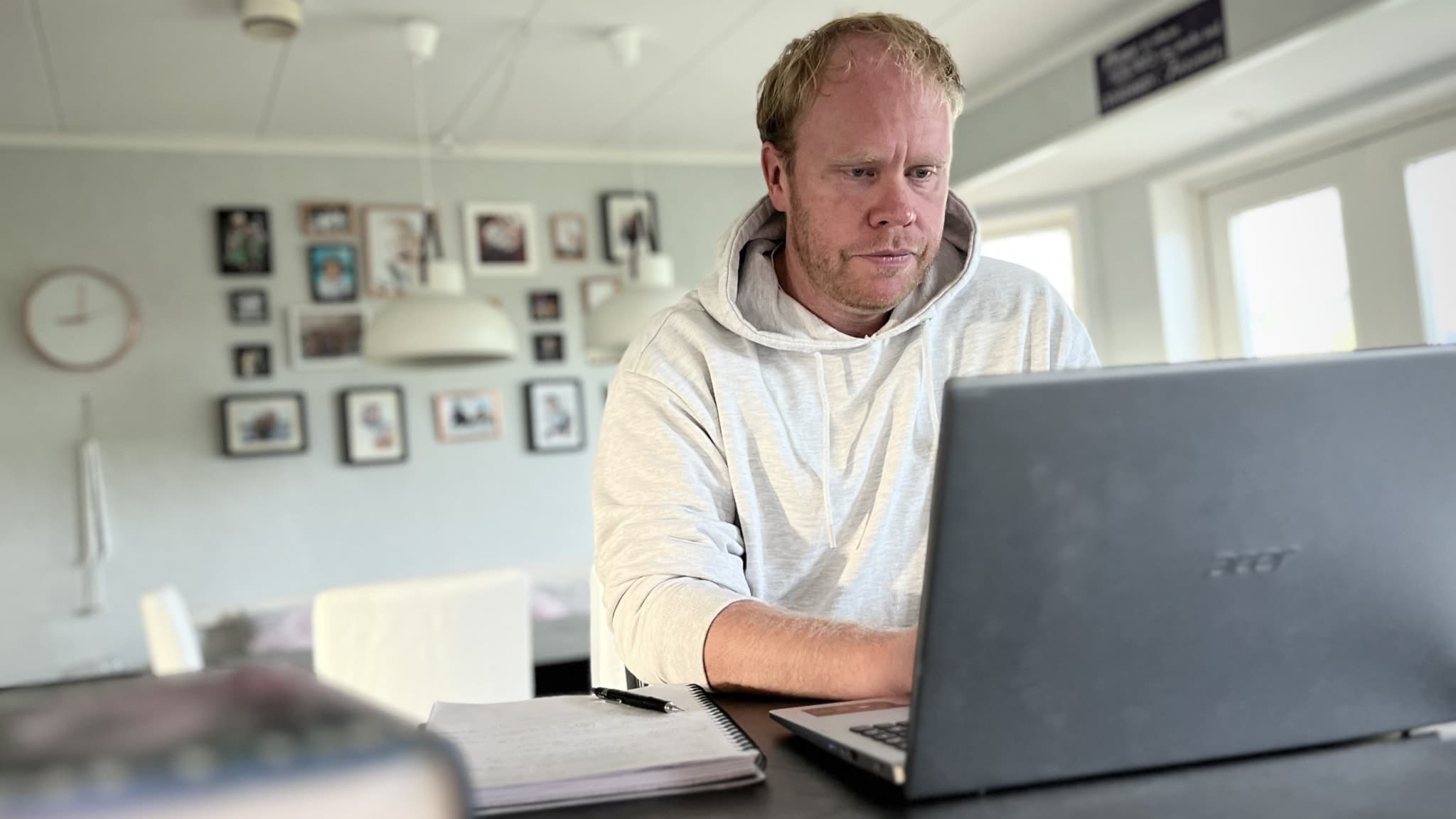 Martin Larsson sitter vid ett bord hemma i Dalarna och studerar på sin bärbara dator, med böcker och anteckningar framför sig. Väggarna bakom honom är prydda med familjefotografier.