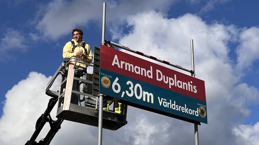 Armand Duplantis står på en skylift bredvid en skylt med texten '6,30 m Världsrekord' och sitt namn, med molnig himmel i bakgrunden. 