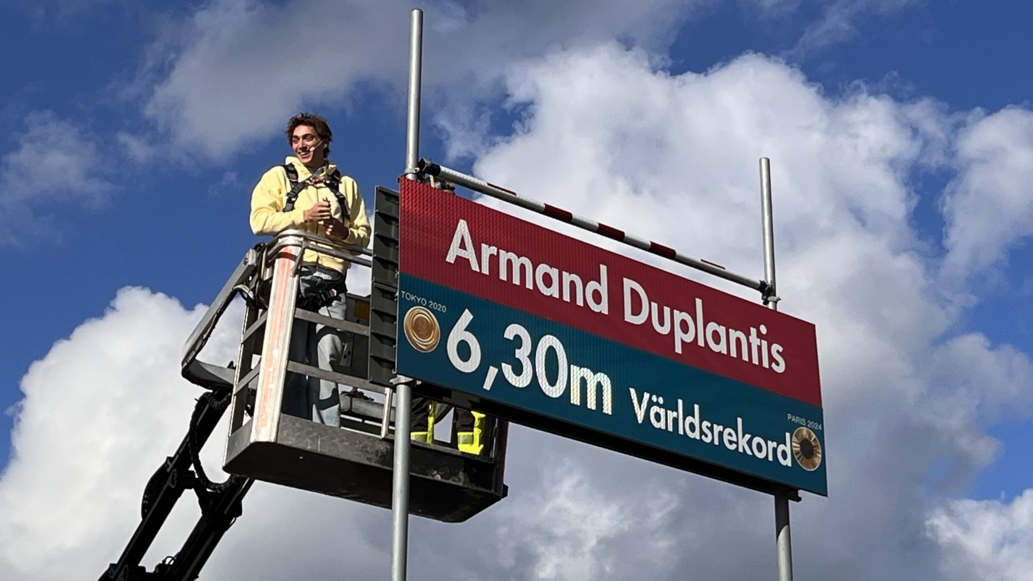 Armand Duplantis står på en skylift bredvid en skylt med texten '6,30 m Världsrekord' och sitt namn, med molnig himmel i bakgrunden. 