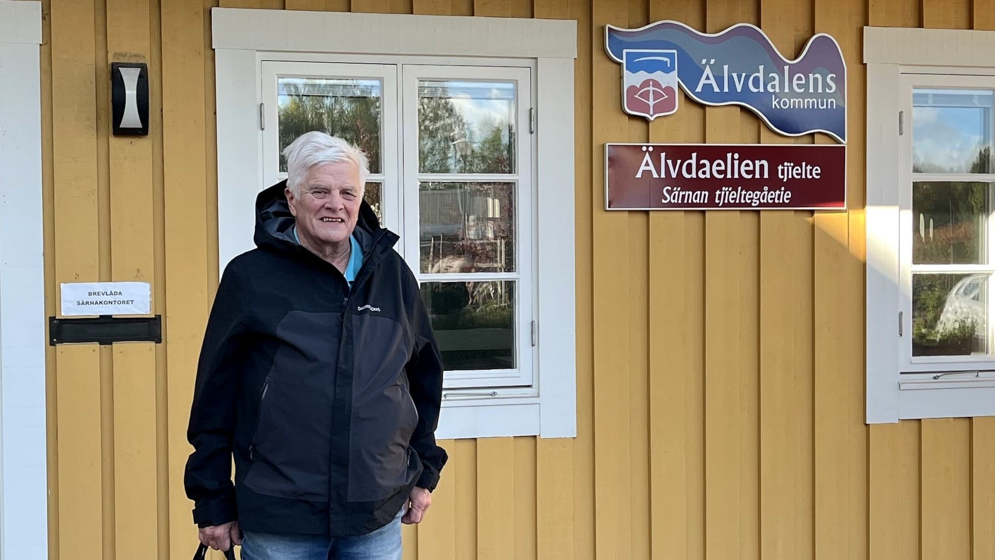 Stig Olsson står framför en gul byggnad med skylten 'Särnakontoret'. Han håller en svart väska och har på sig en mörk jacka. Solen lyser och reflekterar i fönstren.