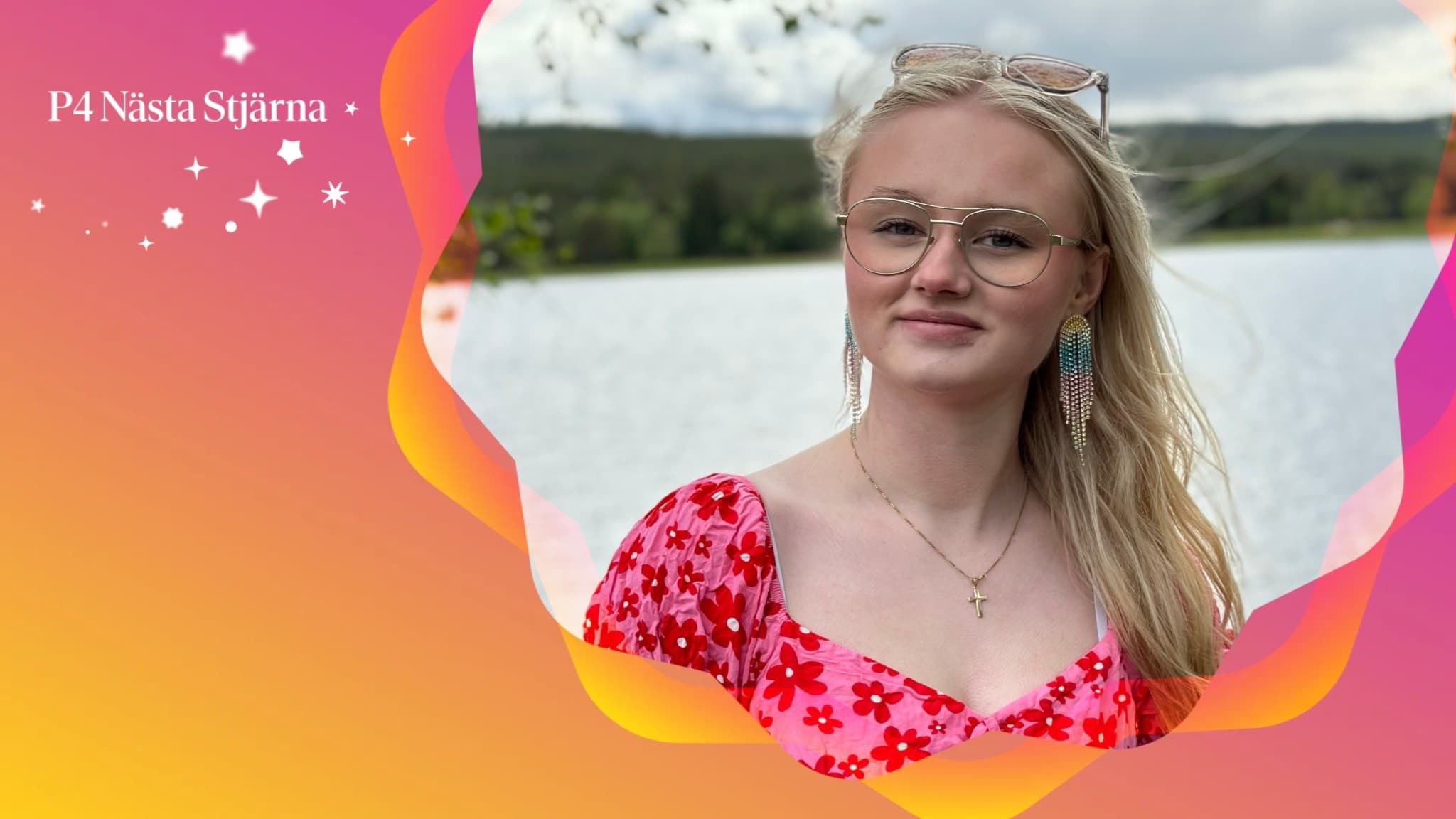 Amelia Lejonclou, finalist i P4 Nästa Stjärna, poserar utomhus framför en sjö med gröna kullar i bakgrunden. Hon bär en röd blommig klänning.