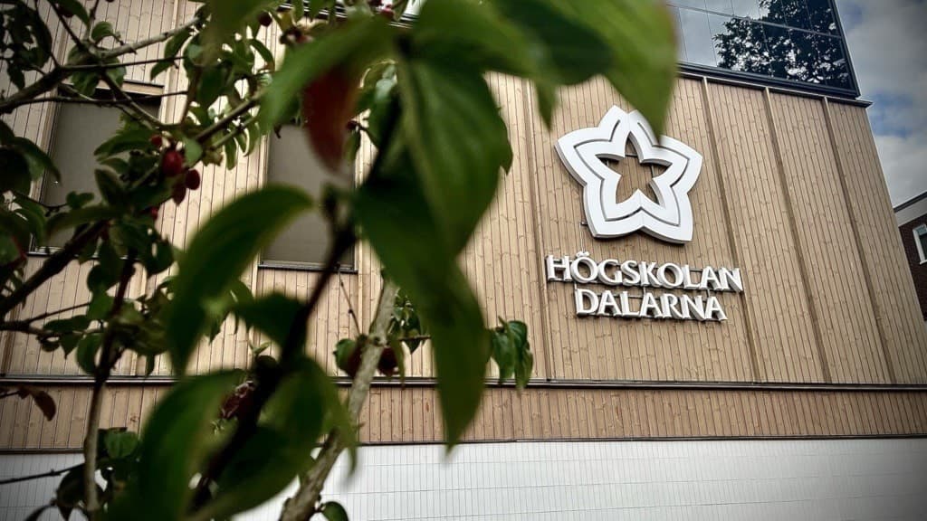 Byggnad med högskolan Dalarnas logotyp synlig på fasaden. I förgrunden skymtar grenar från ett träd, vilket ger ett naturligt inramat perspektiv på den akademiska miljön.