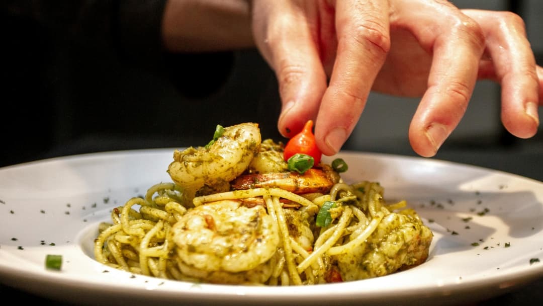 En tallrik med pasta toppad med räkor i grön pesto, dekorerad med en liten röd tomat. En hand placerar tomaten försiktigt på maten, vilket ger en känsla av detaljfokus och gastronomisk precision.