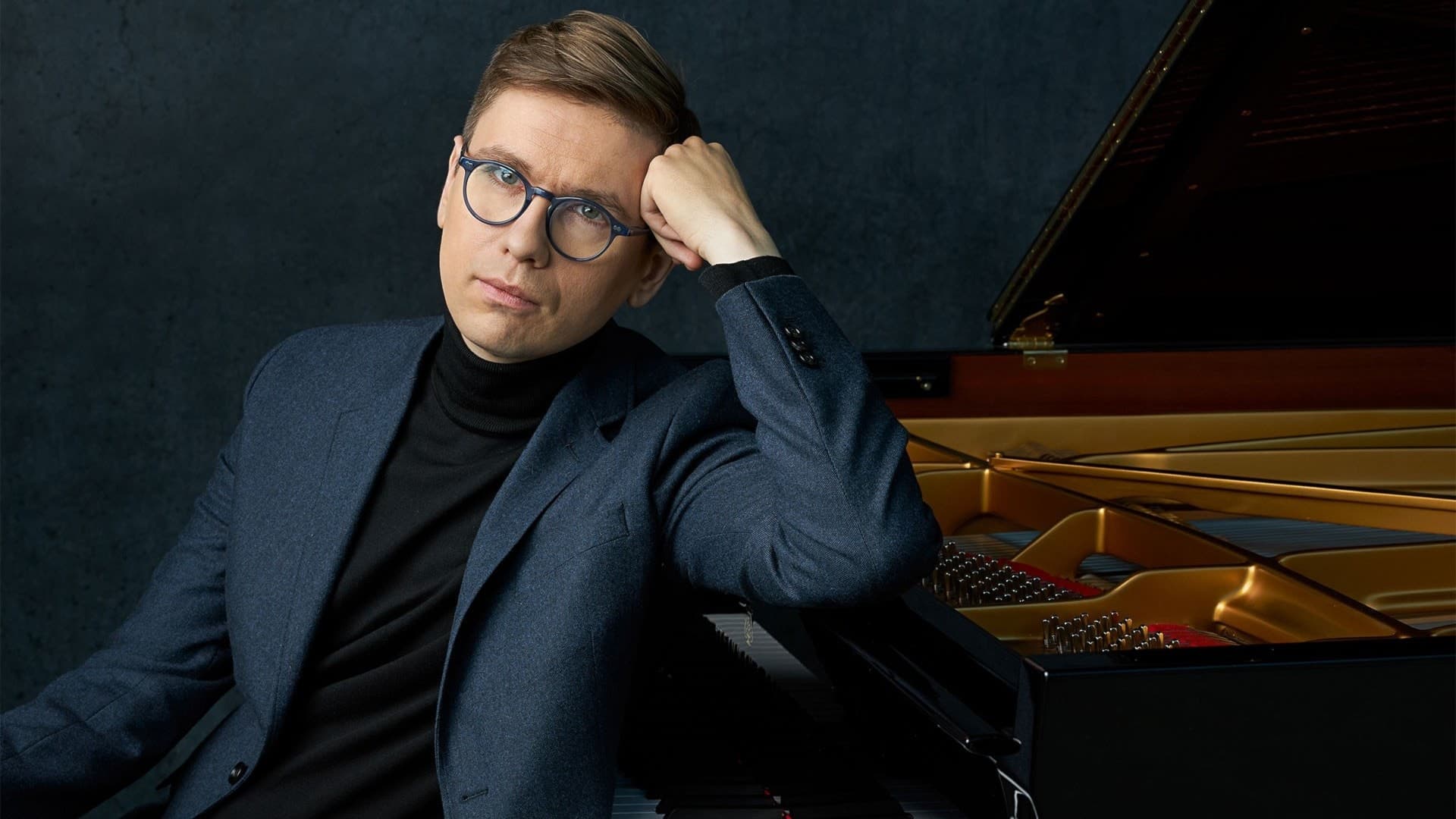 En person i blå kavaj sitter vid ett piano med handen vilande mot huvudet. Pianot är öppet och detaljer av tangentbordet och insidan syns tydligt.