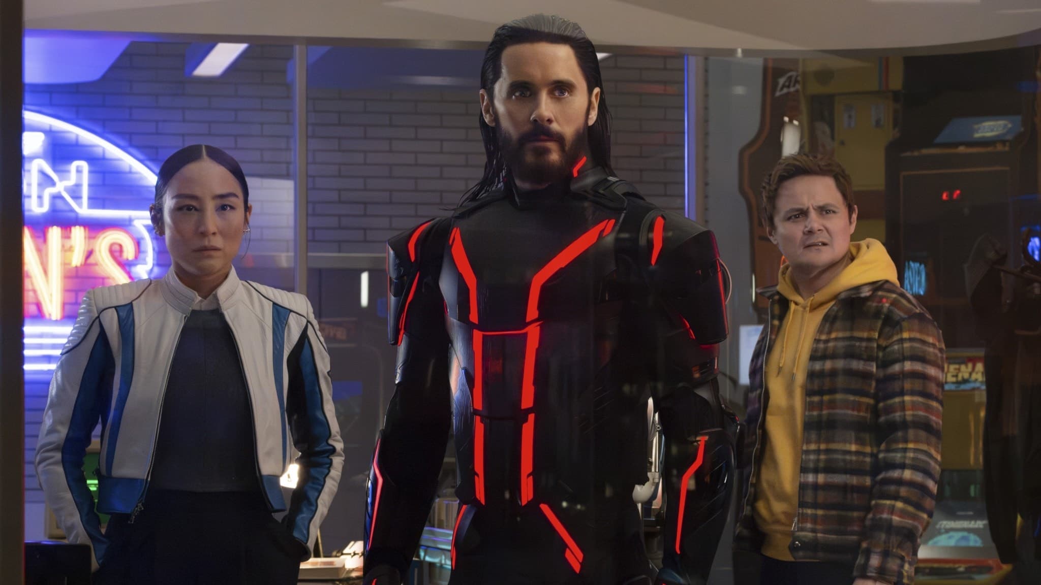 Greta Lee, Jared Leto och Arturo Castro syns i futuristiska kläder i en scen från filmen ”Tron: Ares”, omgiven av neonljus och teknologisk inredning.