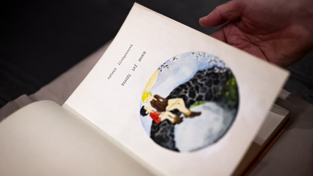 En hand håller en uppslagen bok av Astrid Lindgren, 'Bröderna Lejonhjärta'. Boken visar en illustration av två karaktärer på en klippa. Boken är en nyutgåva med fokus på yttrandefrihet.