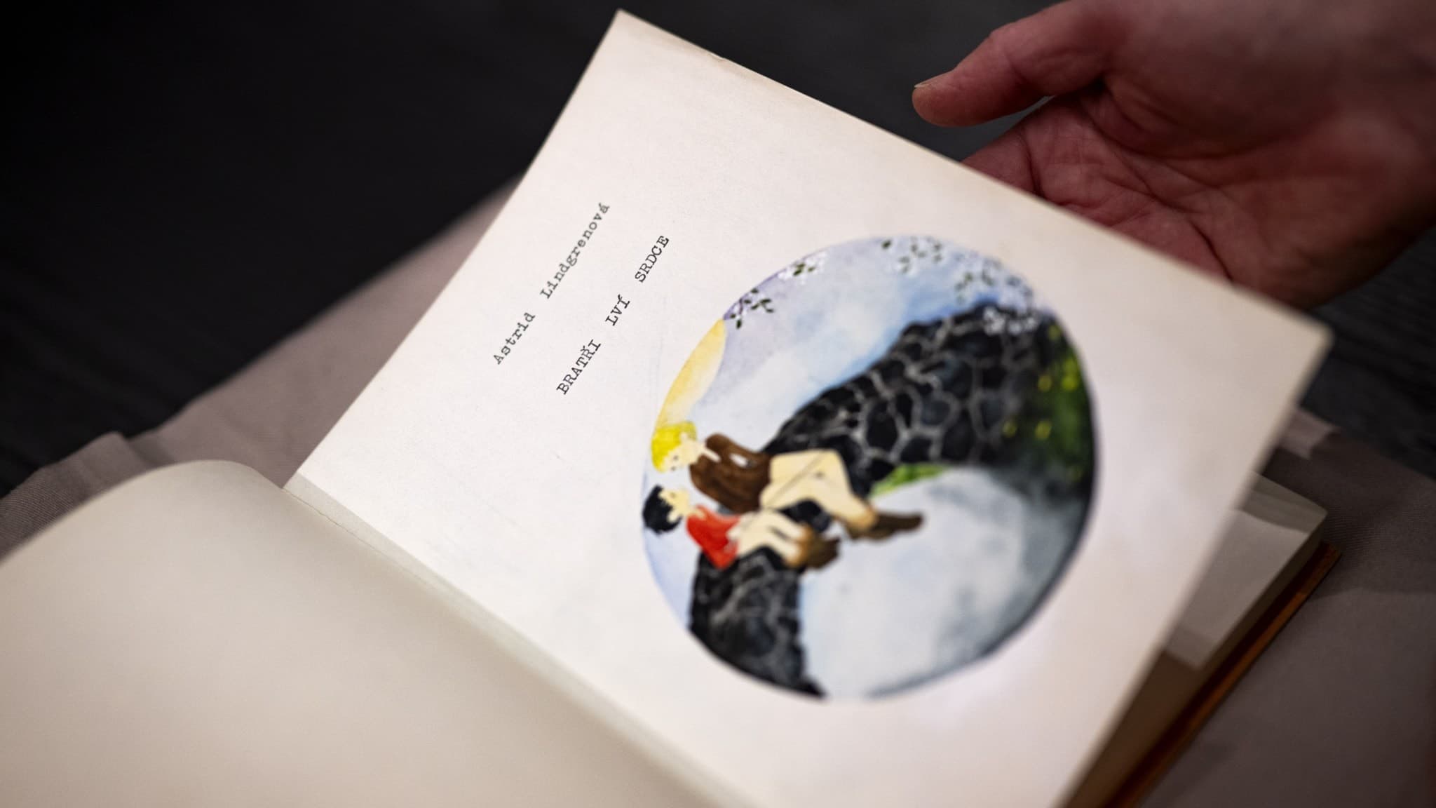 En hand håller en uppslagen bok av Astrid Lindgren, 'Bröderna Lejonhjärta'. Boken visar en illustration av två karaktärer på en klippa. Boken är en nyutgåva med fokus på yttrandefrihet.