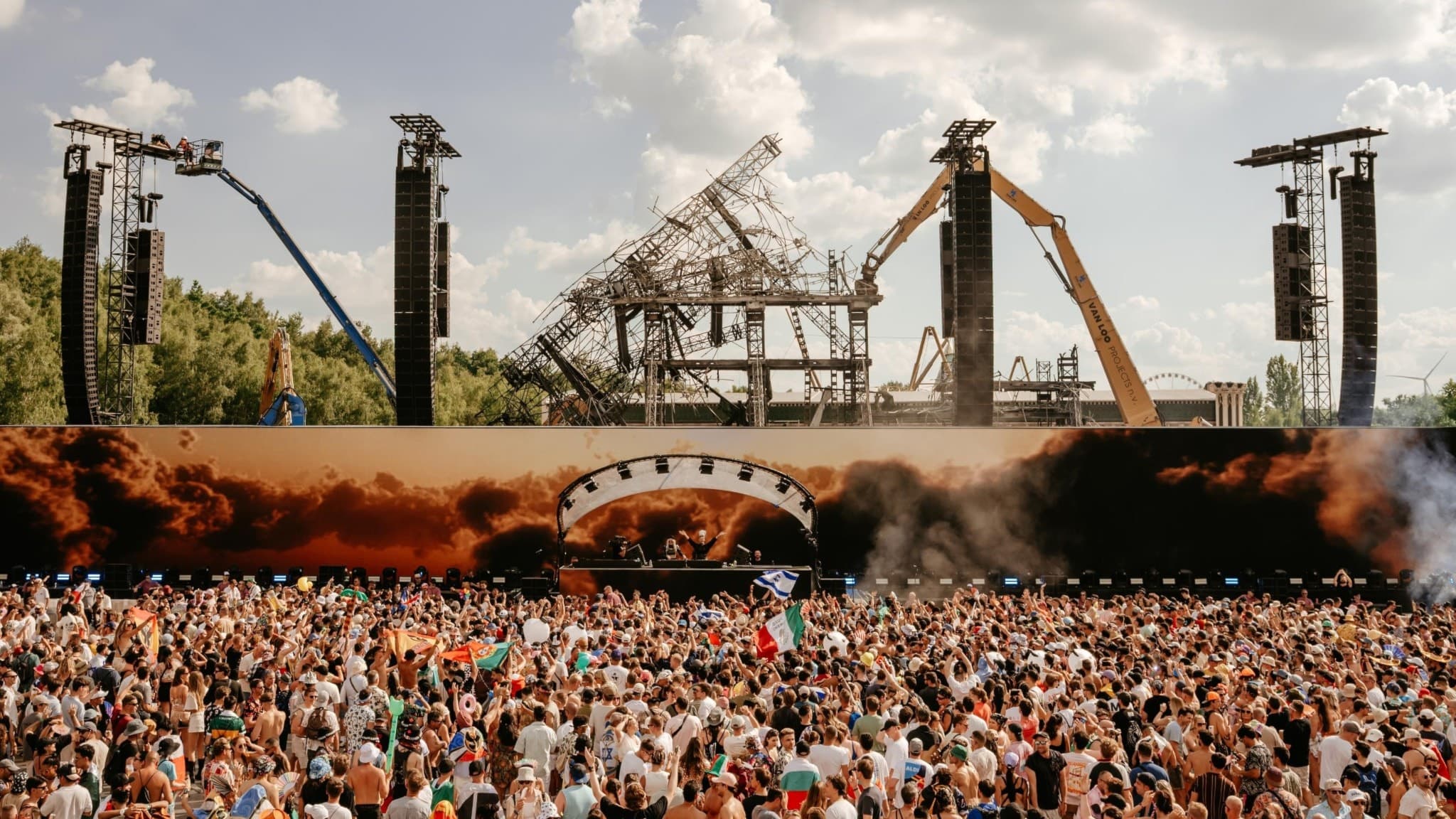 På bilden syns ett foto av den nya stora scenen på Tomorrowland 2025 efter att den först abrunnit ned. Ett stort folkhav av punlik står framför och i bakgrunden syns resterna av den gamla secnen och en grävskopa.