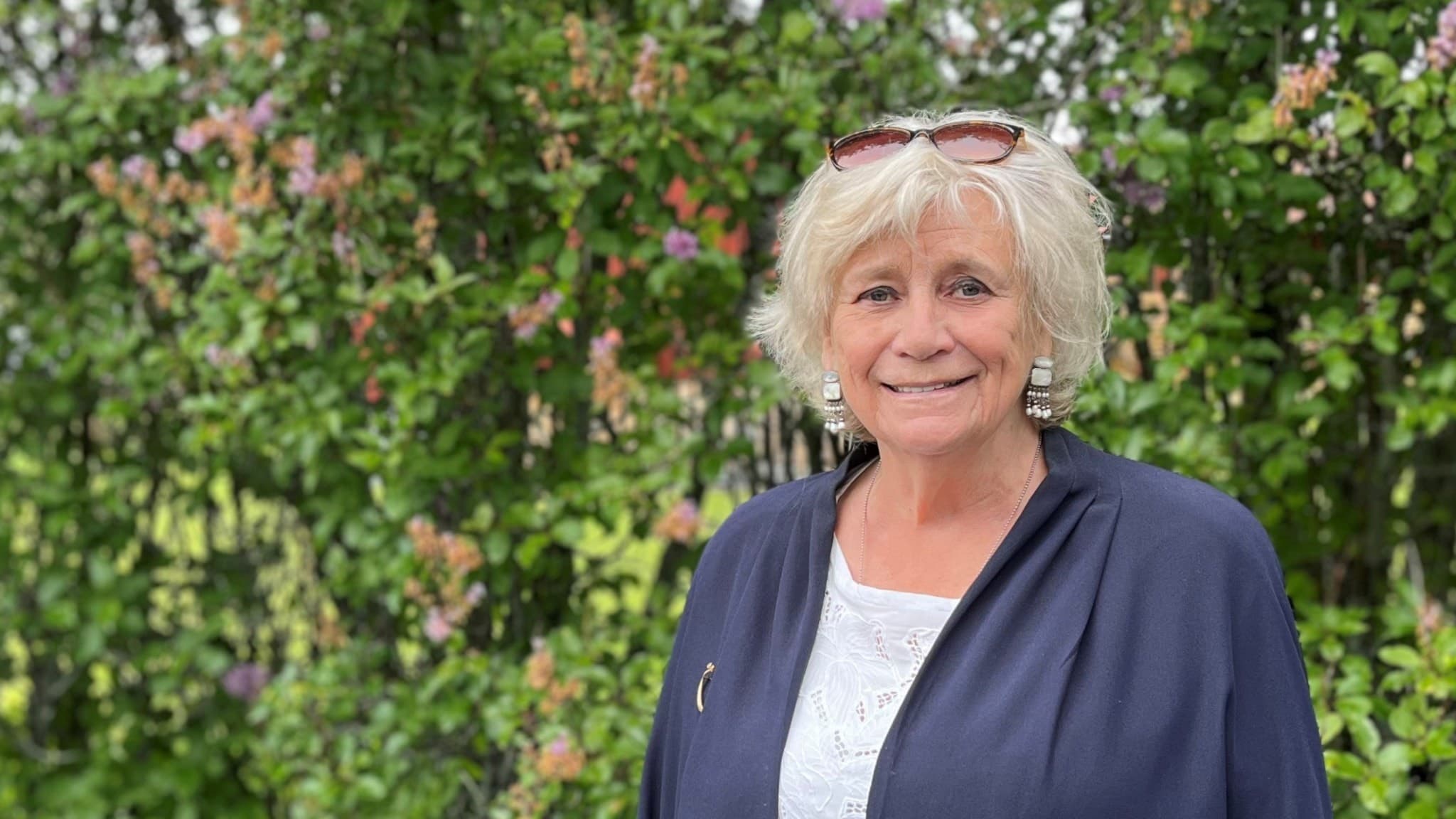 En sommarklädd kvinna med solglasögon uppskjutna i det grålockiga håret ler in i kameran