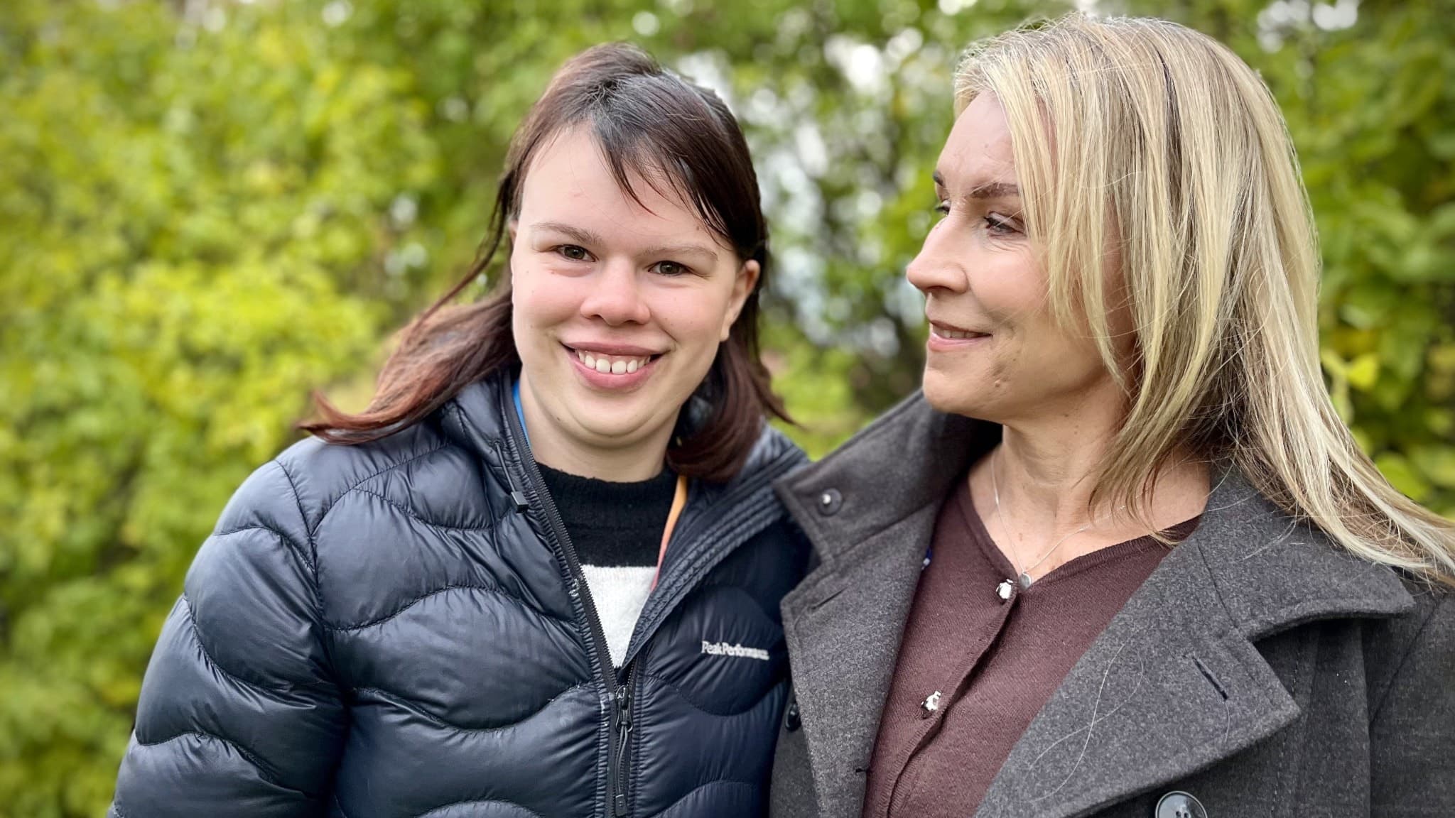 Två kvinnor står utomhus framför gröna träd, klädda i mörka jackor. Den ena har långt blont hår och den andra mörkt hår. 