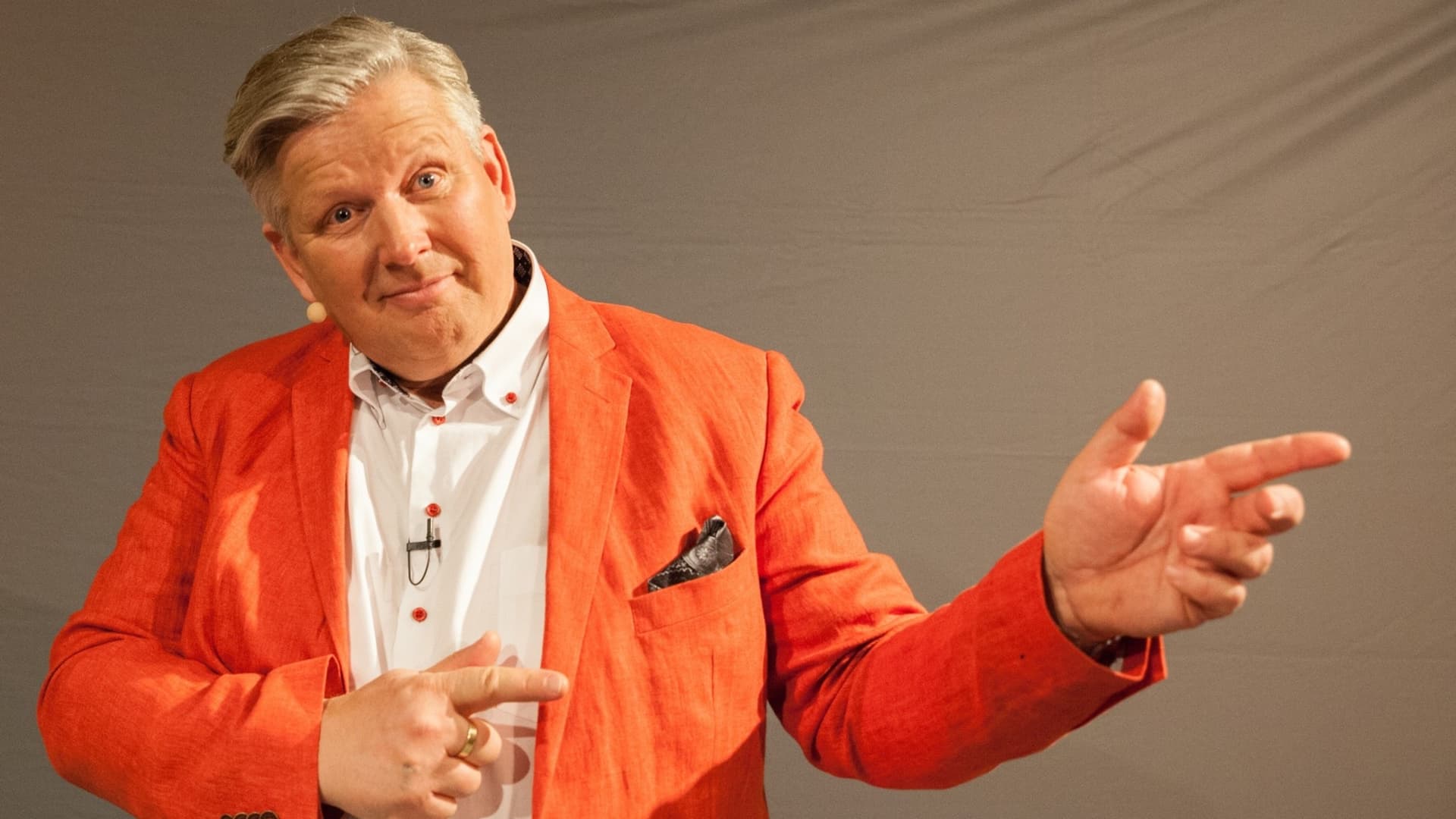 Mats Danielsson, klädd i en orange kavaj och vit skjorta, gestikulerar med händerna framför en neutral bakgrund. Han är känd som expert på vett och etikett.