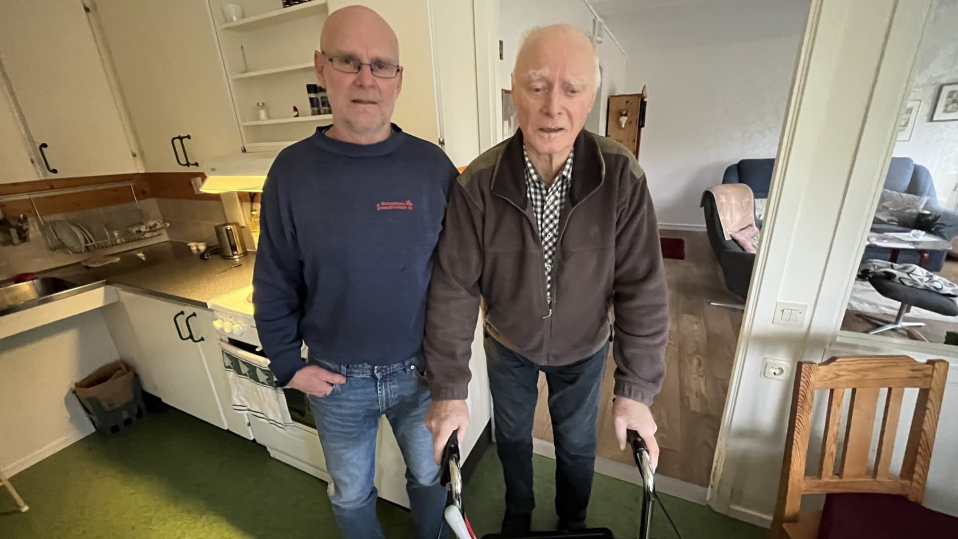 Harald Forsberg står bredvid sin morbror Bengt Kärnlund som använder en rollator. Bilden är tagen i ett hem med köksinredning i bakgrunden och visar påverkan av bristande vårdkommunikation.