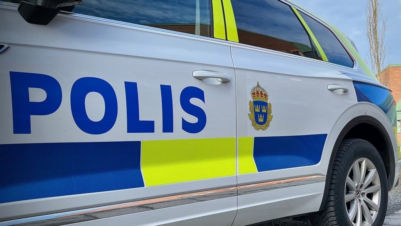 En målad polisbil i vitt, blått och gult framför en stor tegelbyggnad med grönt tak. 