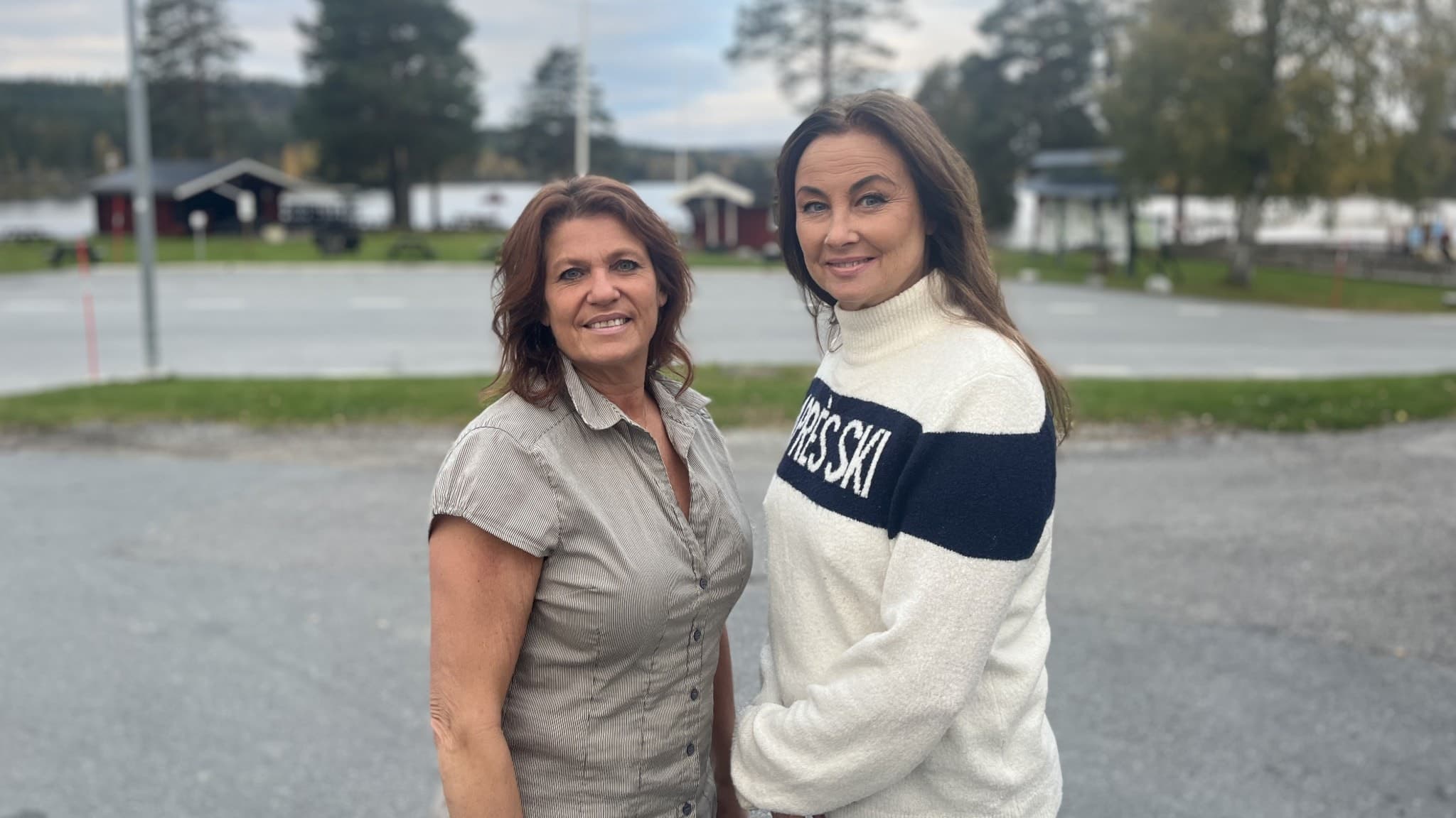 Carola Engström och Ingela Frimert står utomhus vid en asfalterad plats med träd, sjö och små röda stugor i bakgrunden. De är klädda i vardagliga kläder och ser in i kameran.