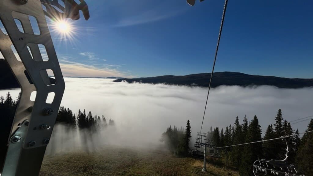 Skidlift med utsikt över dimtäckt dal och solbelysta berg i bakgrunden. Skogsområde täcker landskapet, med klar blå himmel ovanför.