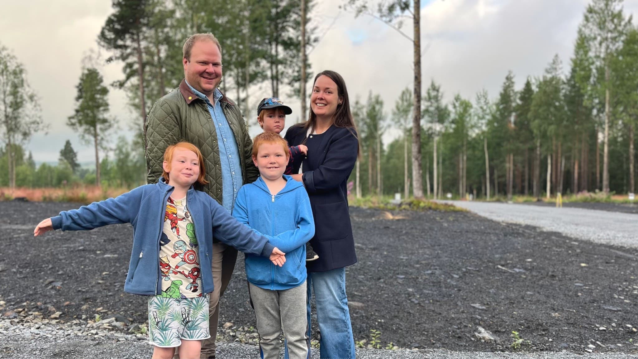 Mattias och Linnea Barkenfelt står med sina barn på en grusväg omgiven av skog. Familjen ler och barnen ser glada ut, med höga träd och en molnig himmel i bakgrunden.