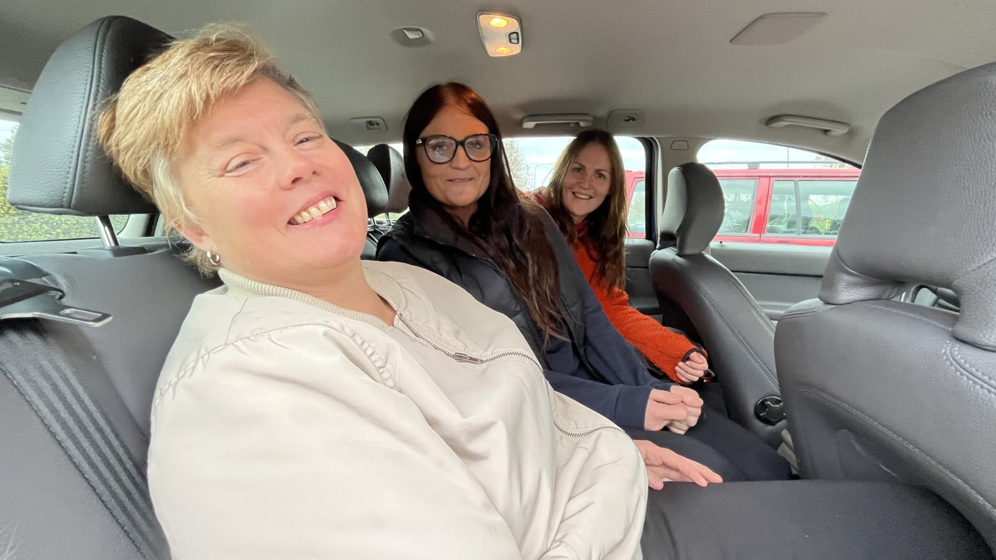 Ellinor Ljung, Erica Tengström och Jenny Carlzon sitter i baksätet på en bil. Ljung är klädd i ljus tröja, Tengström i mörka kläder och Carlzon i orange jacka. En röd bil syns utanför fönstret.
