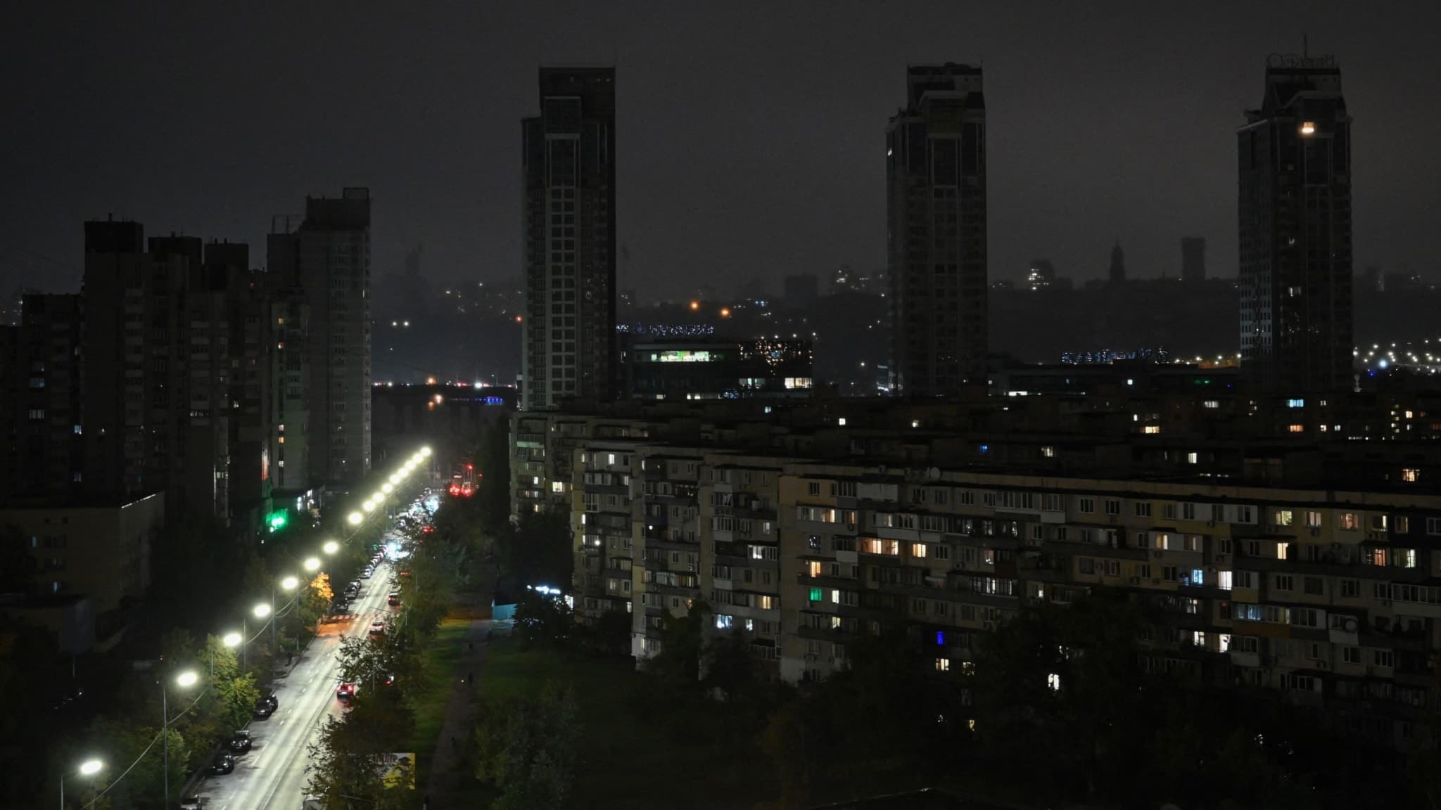 Mörklagda bostadshus vid Dniproflodens östra strand i Kyiv under ett strömavbrott orsakat av ryska missil- och drönarattacker, med en svagt upplyst gata i förgrunden.