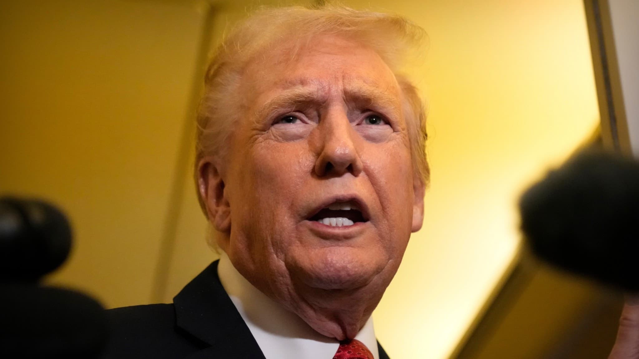 USA:s president Donald Trump iförd kostym och röd slips, talar till reportrar ombord på Air Force One, med mikrofoner synliga i förgrunden.