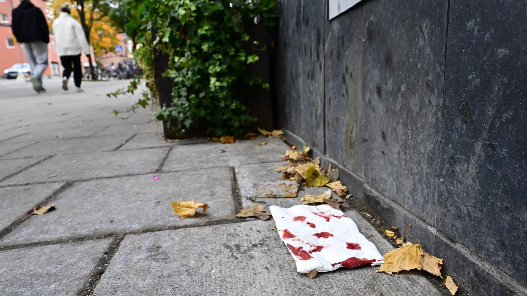 Blodfläckad näsduk ligger på trottoaren vid brottsplatsen för en skottlossning i centrala Gävle. Två personer går i bakgrunden nära butiker och restauranger. Löv täcker delar av marken.
