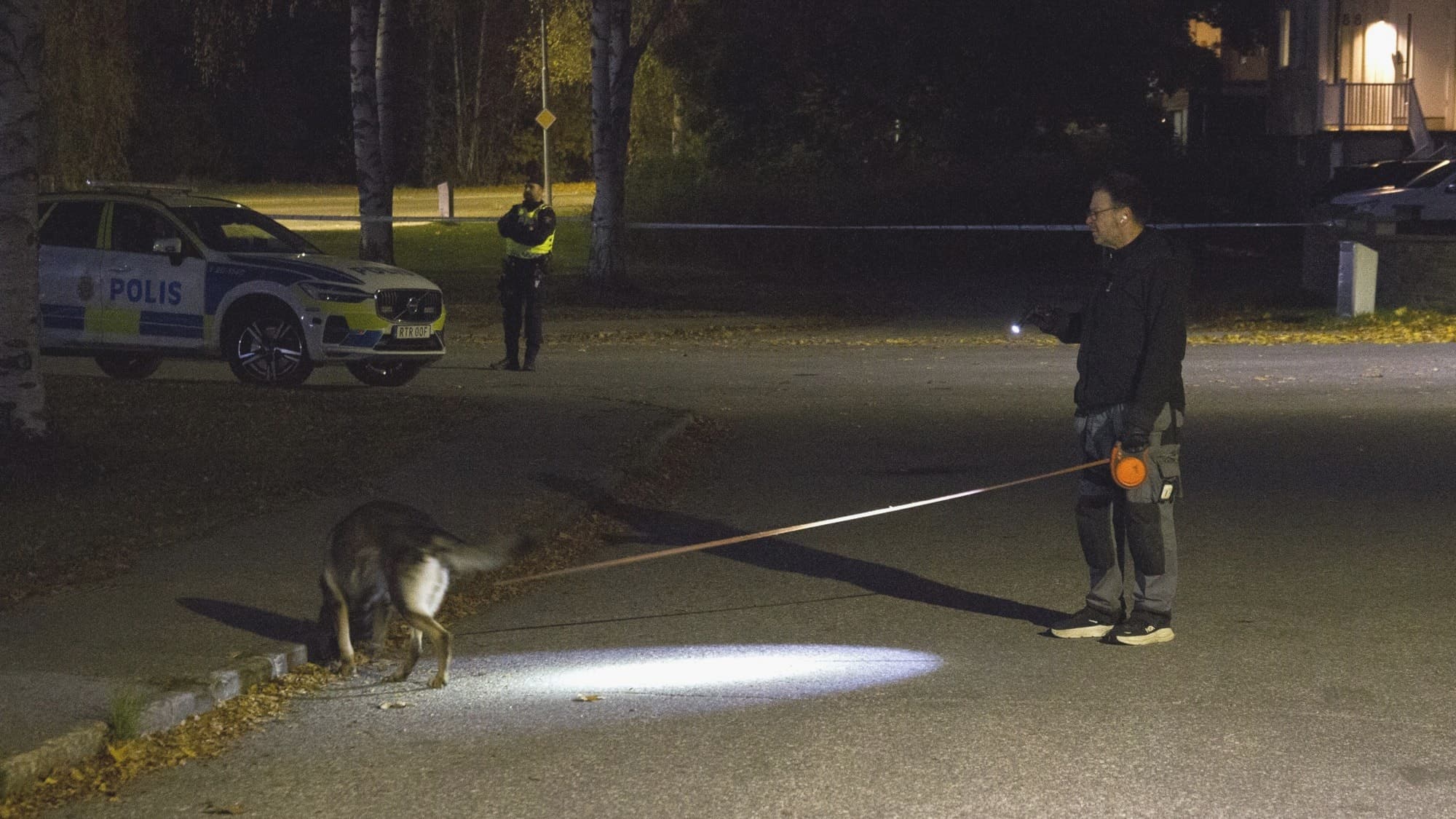 En polis och en spårhund söker efter ledtrådar på en avspärrad plats under natten. En polisbil står i bakgrunden, och en annan polis vaktar området.