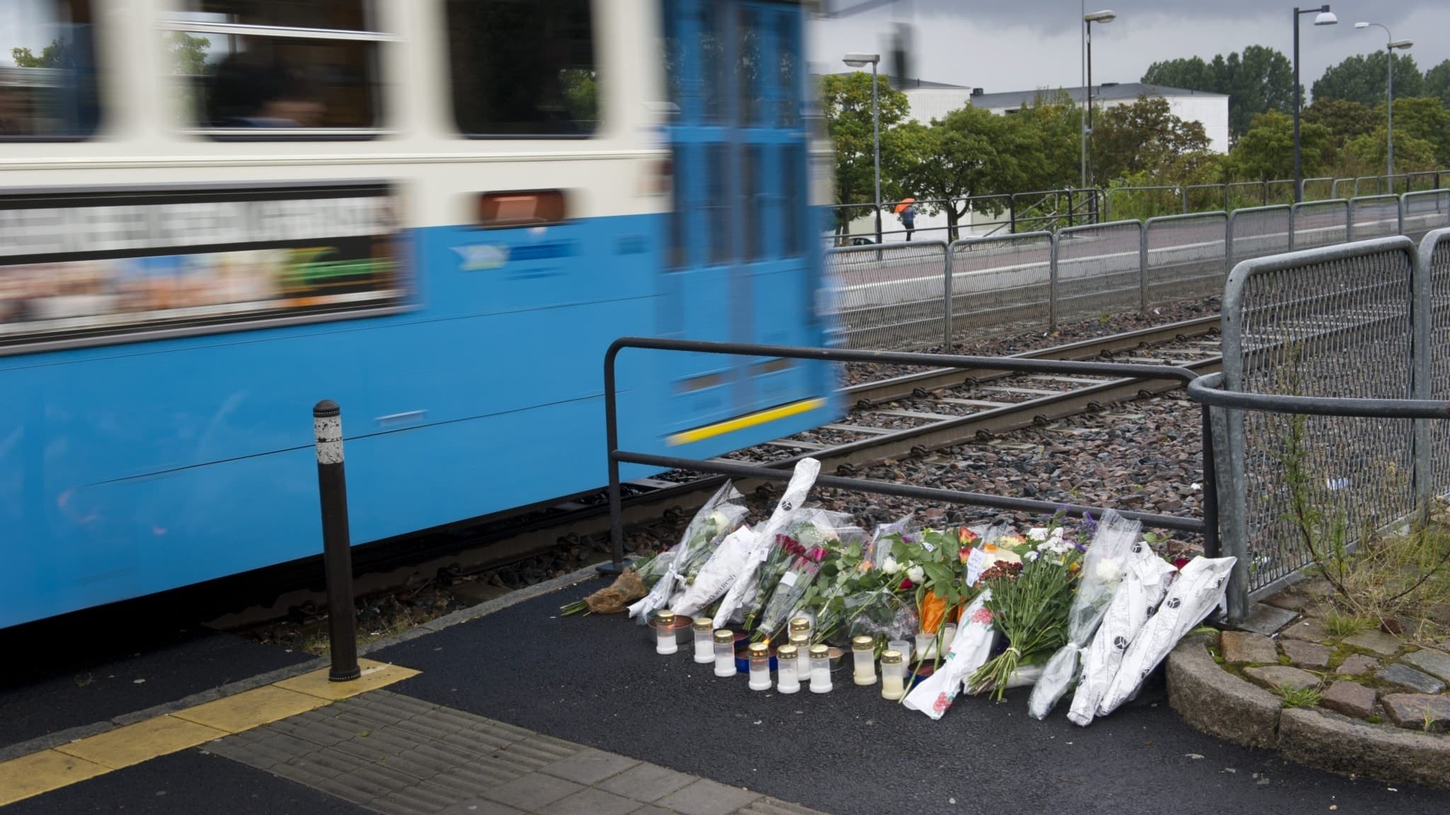 Blommor och ljus ligger vid Briljantgatans spårvagnshållplats i Göteborg, där en 26-årig man sköts till döds. En spårvagn passerar platsen, omgiven av stängsel och gråa skyar.