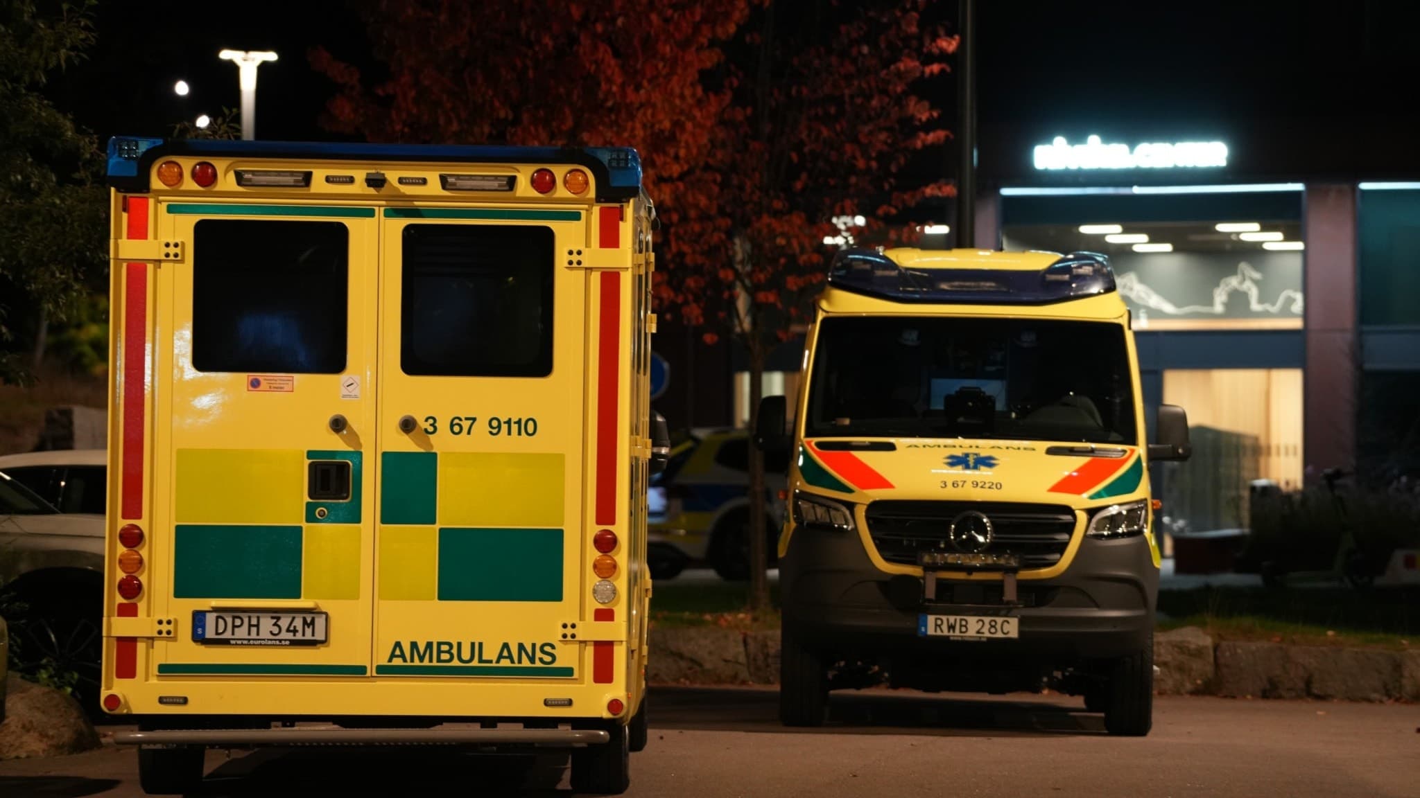 Två ambulanser står parkerade i ett upplyst område under kvällen, med ett träd i höstfärger och en byggnad i bakgrunden.
