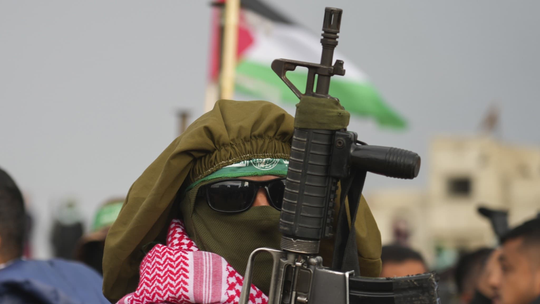 En beväpnad Hamassoldat med ansiktet täckt och ett gevär i händerna står framför en palestinsk flagga i Gaza.