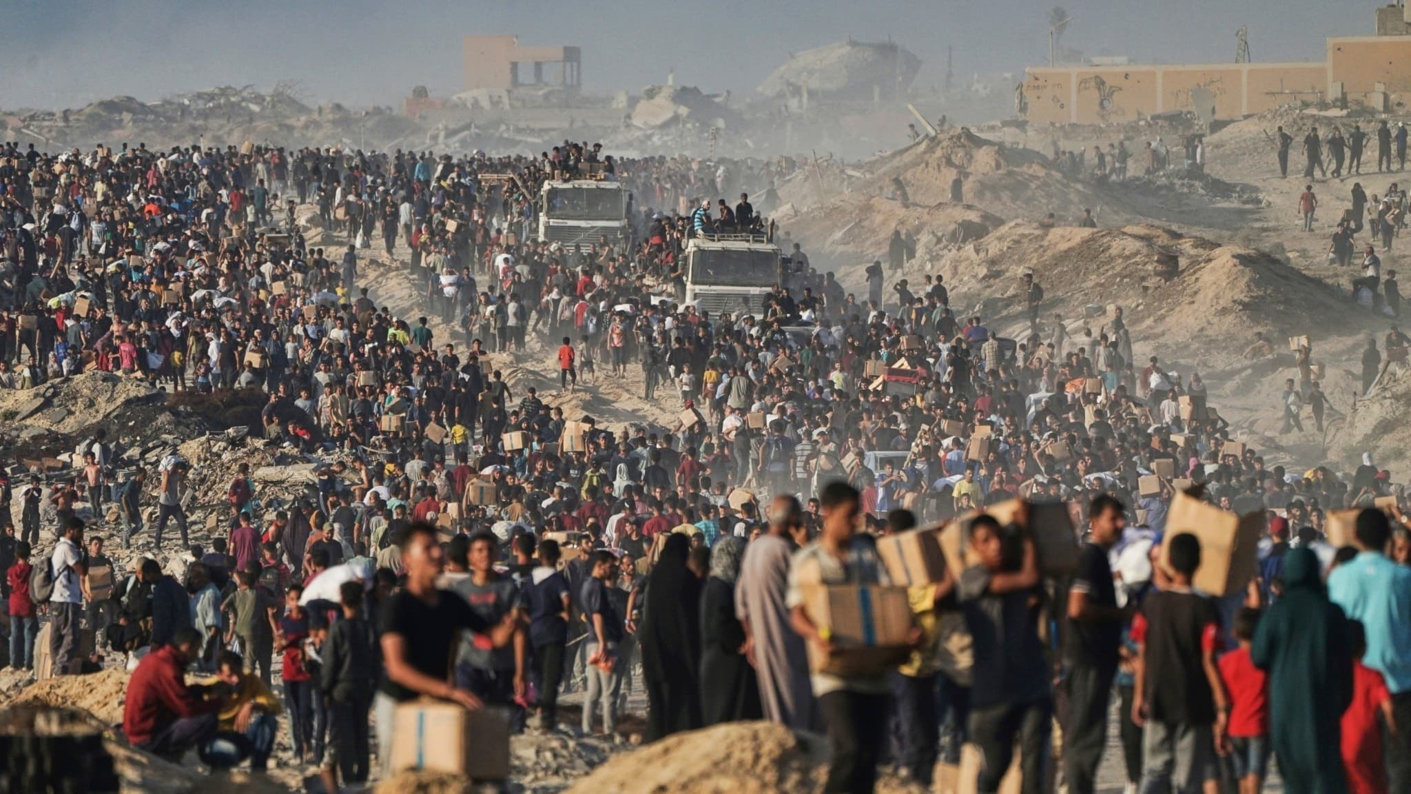 Hundratals människor samlas i norra Gaza för att ta emot mat och humanitär hjälp från ett FN-konvoj. Två lastbilar syns i bakgrunden, omgivna av damm och rörelse.