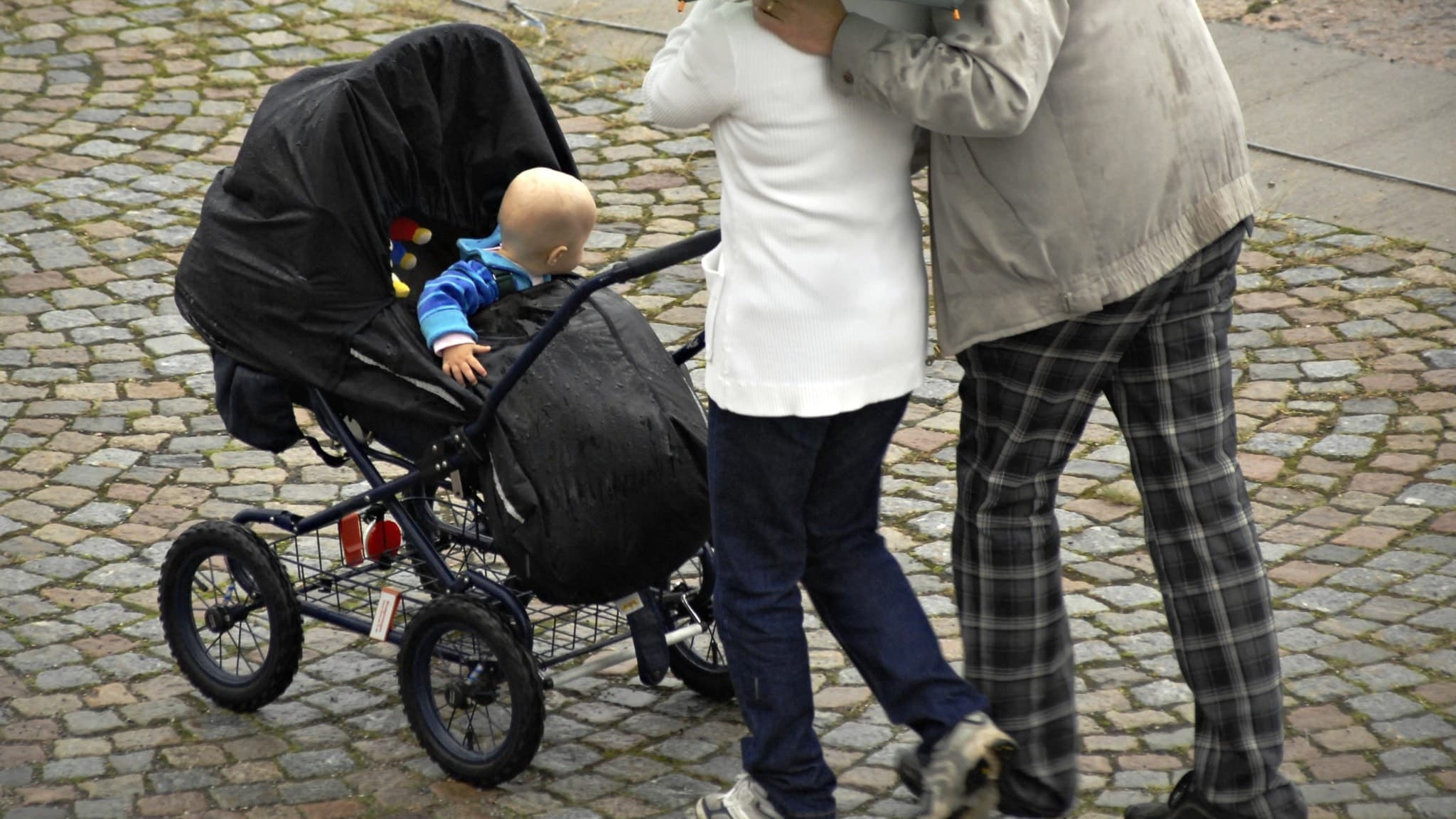 Barnfamilj promenerar på en regnig kullerstensgata i Stockholm. Föräldrarna håller ett grönt paraply och en barnvagn med regnskydd, medan ett litet barn tittar fram från vagnen.