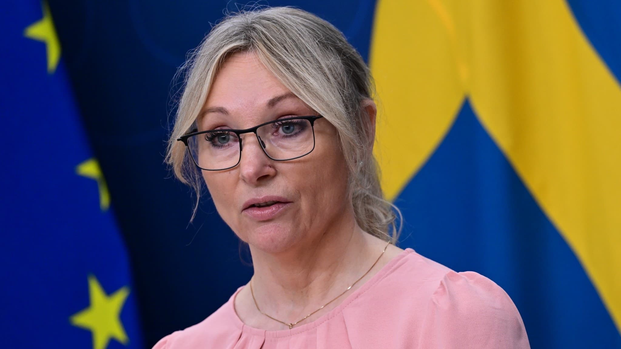 Jessica Stegrud, Sverigedemokraternas socialpolitiska talesperson, talar under en pressträff om välfärdsbrottslighet. I bakgrunden syns EU-flaggan och Sveriges flagga.
