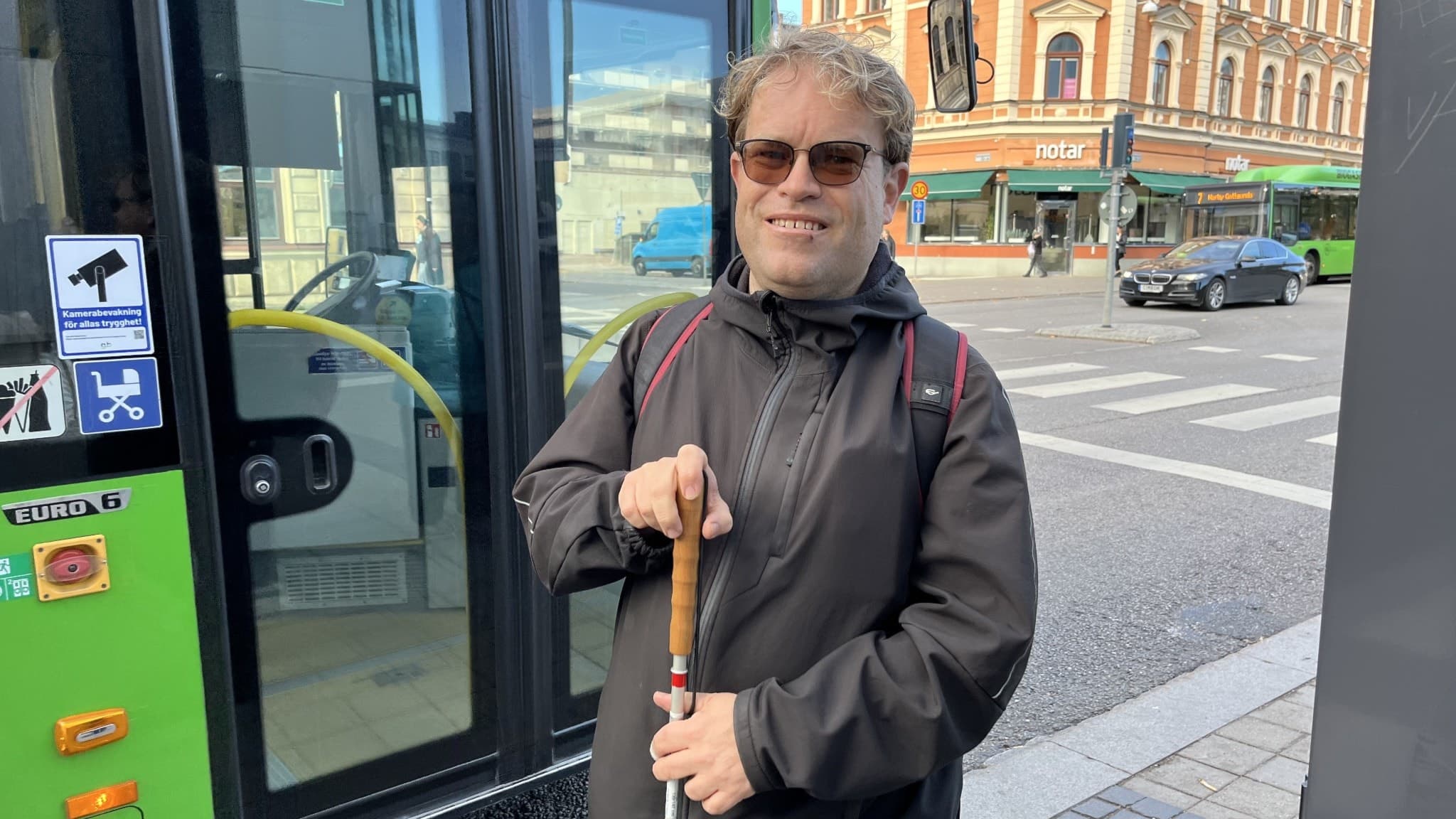 Måns Högström står vid en grön stadsbuss med en vit käpp i handen på en trottoar. I bakgrunden syns en äldre byggnad och en stadsmiljö med trafik.